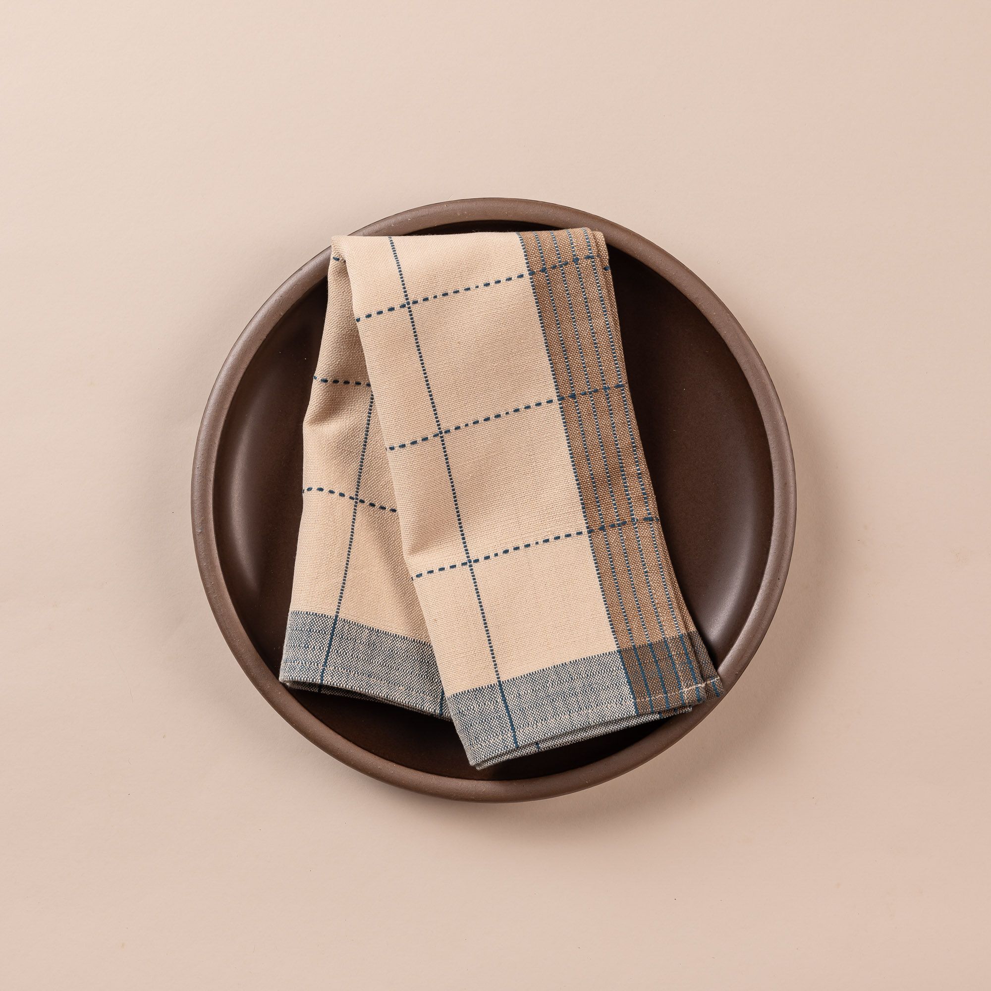 A natural folded napkin sits on a deep brown dinner plate. The napkin is designed with turquoise gridlines with light blue and tan striped edging.