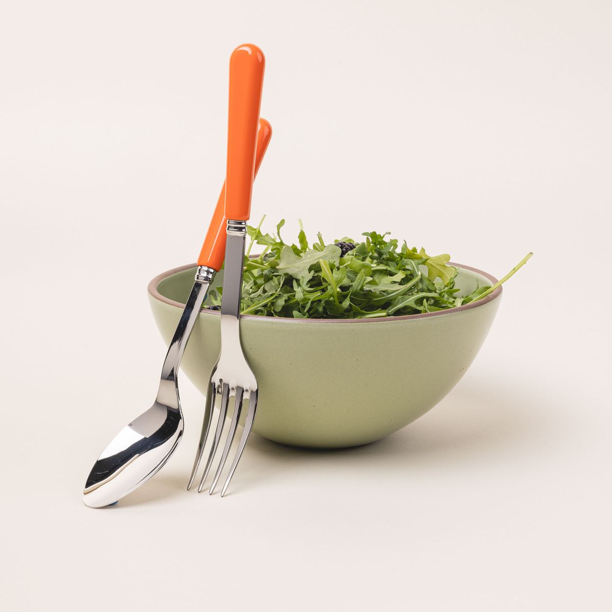 A serving size spoon and fork in a shiny steel with a bold orange handle sits against a calming sage green ceramic bowl filled with salad.