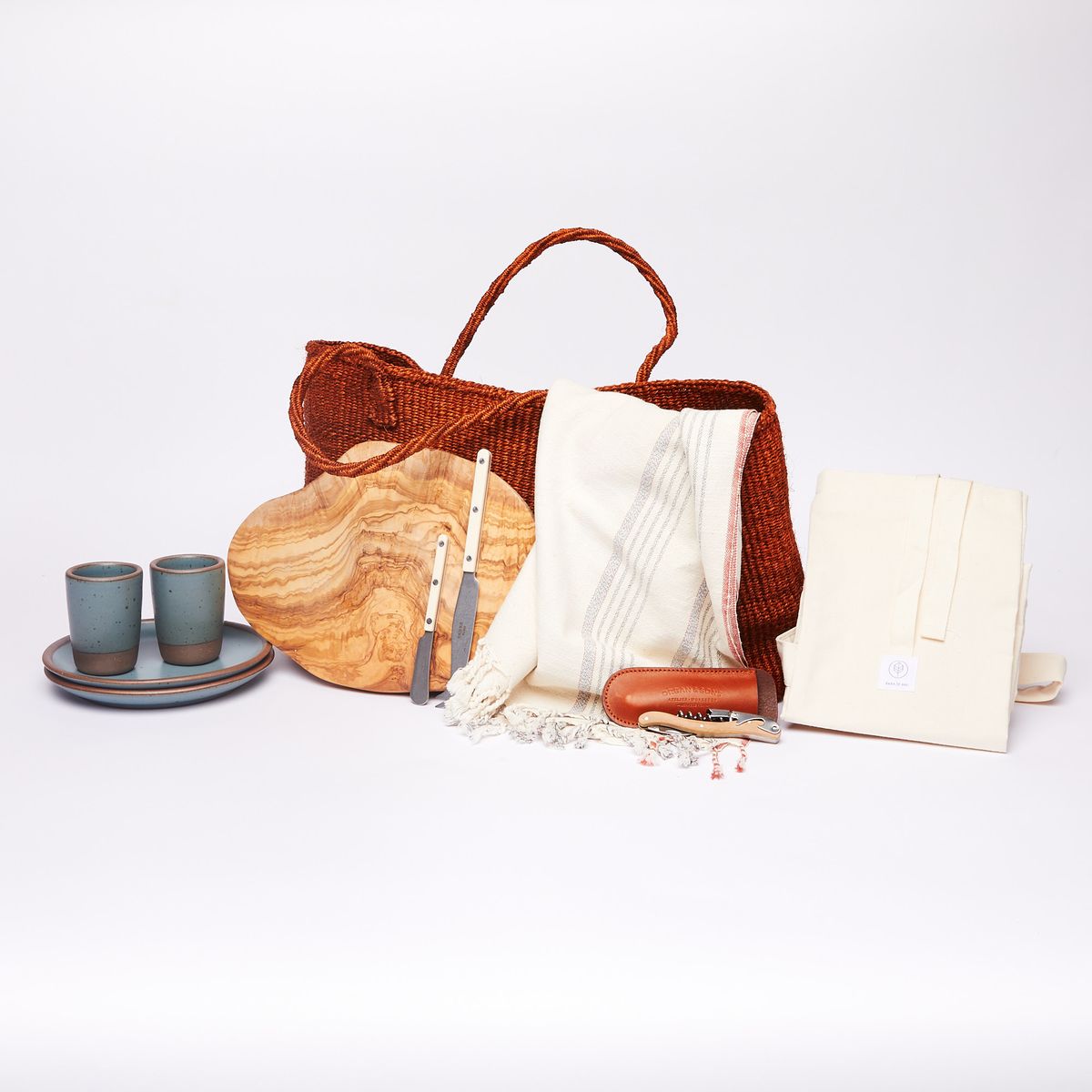 From left to right: Two turquoise juice cups on two turquoise side plates, an rounded olivewood cutting board, a large rust-colored woven tote, two cheese spreader, a striped turkish towel, a wine key with leather sheath, a folded cotton baguette bag