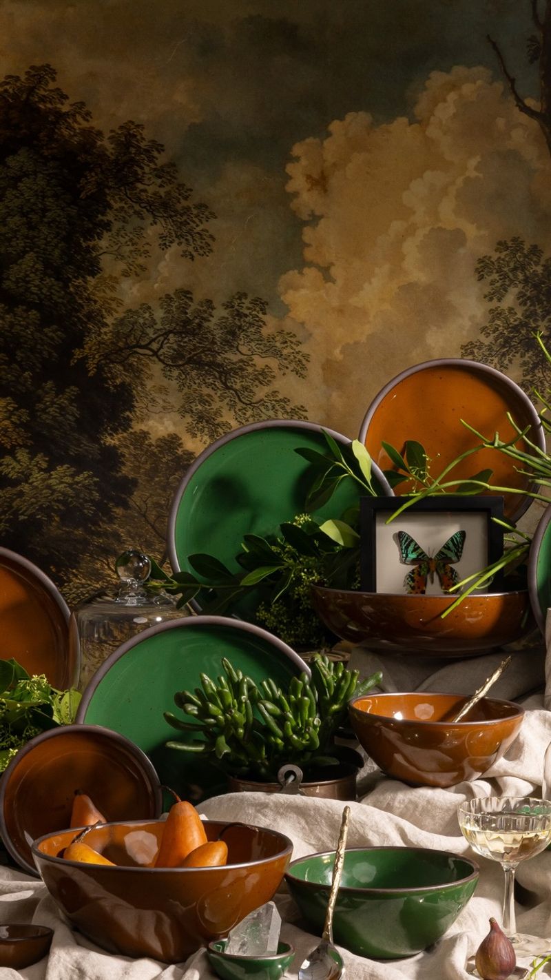 Table with glossy green and brown ceramic plates, candles, crystals, plants, and framed insects against a forest mural backdrop.