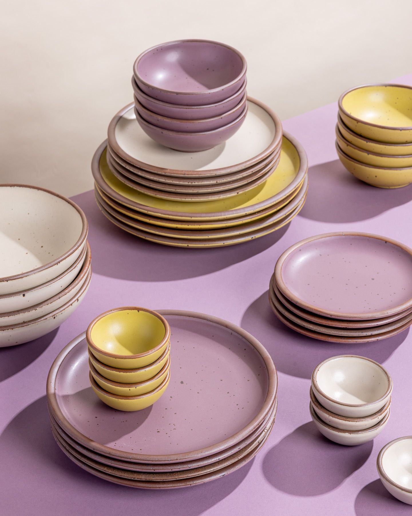Stacks of ceramic plates and bowls in sets of 4, in colors including zesty yellow, soft mauve purple, and warm off-white.