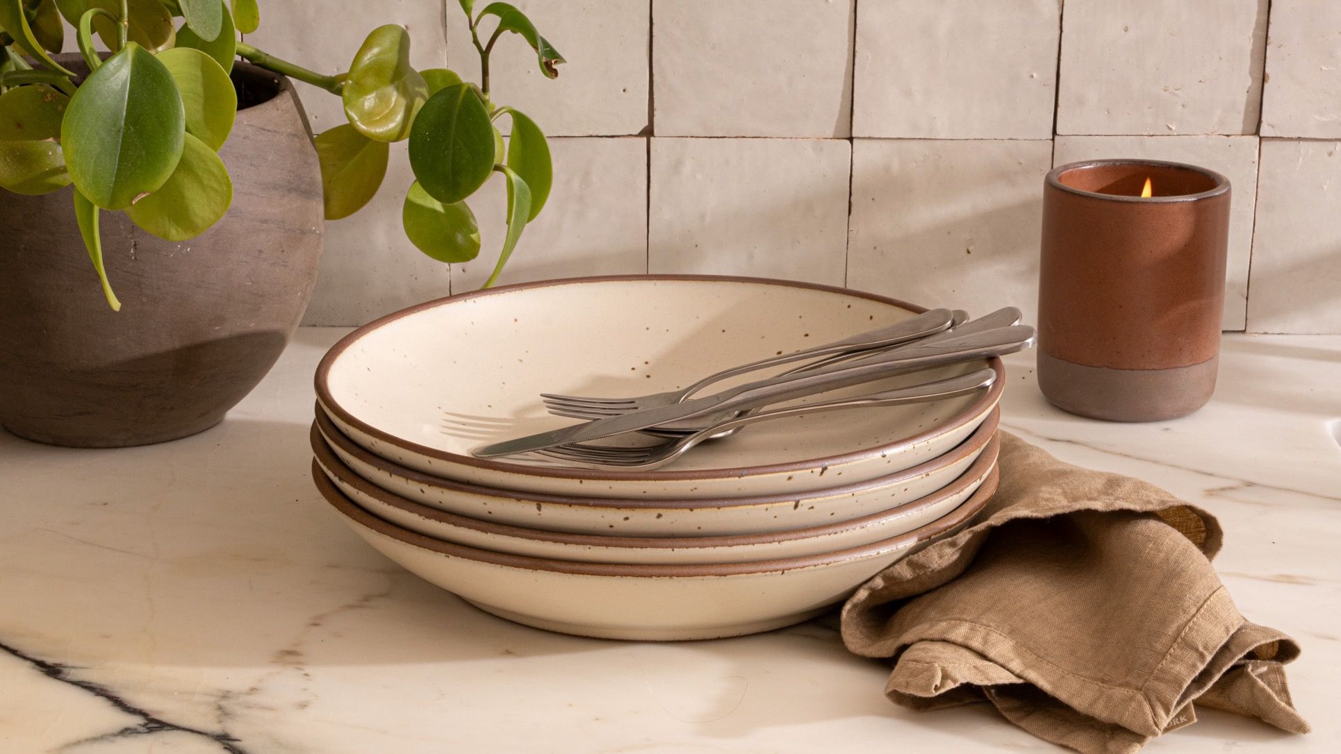 A stack of warm off-white coupe plates with silverware, a tan napkin, a candle, and a potted plant on a countertop.