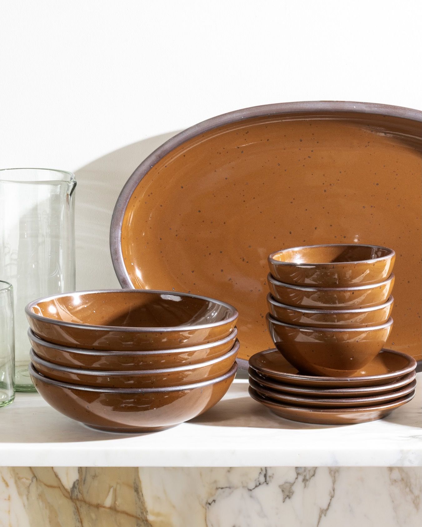 Stacked glossy caramel brown ceramic bowls and plates with a large oval platter on a marble shelf.