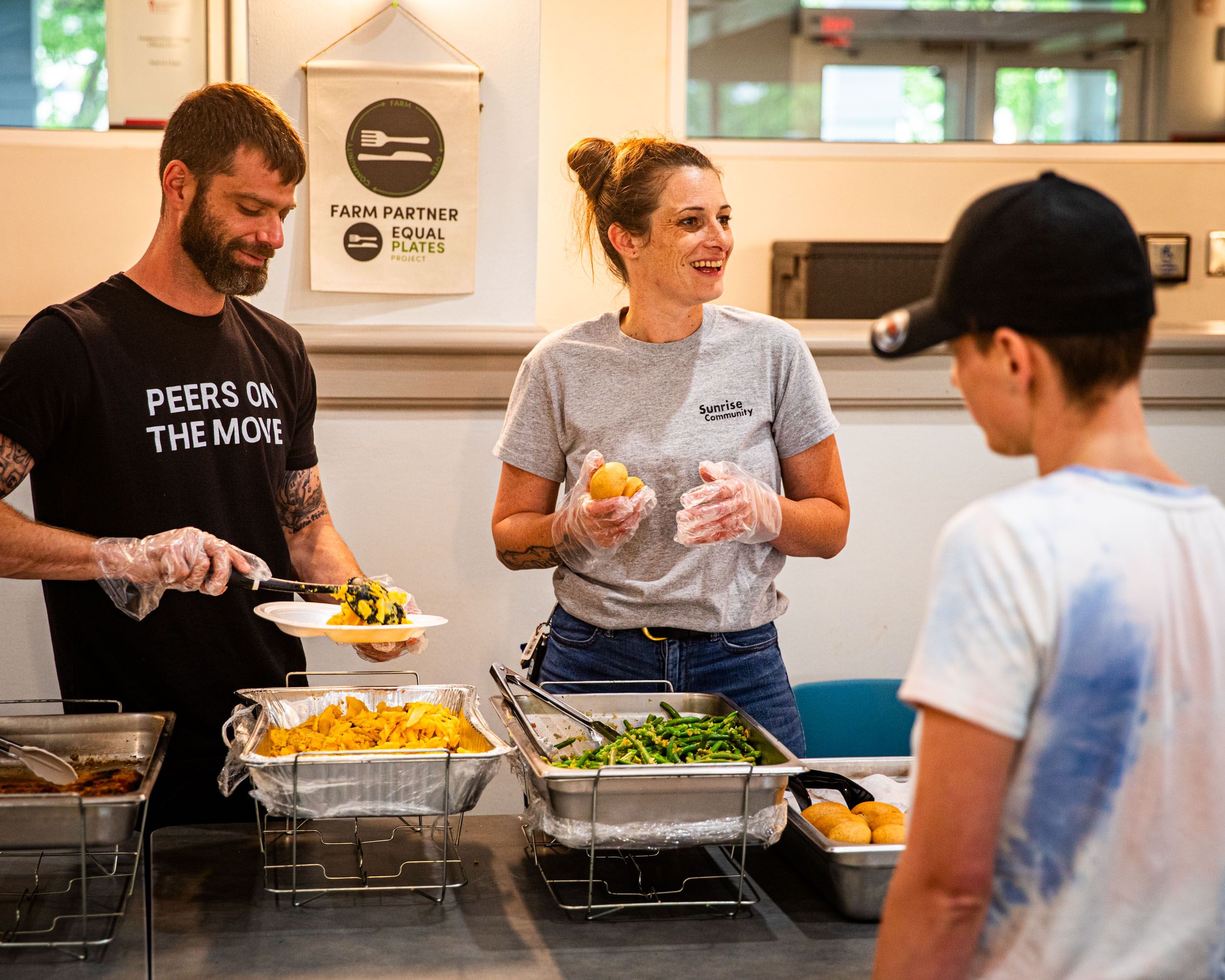 Two people are standing behind a food line, serving foods on plates for someone in line.