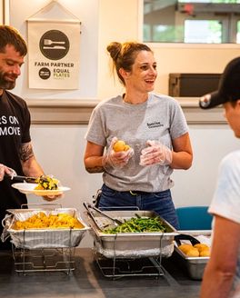 Two people are standing behind a food line, serving foods on plates for someone in line.