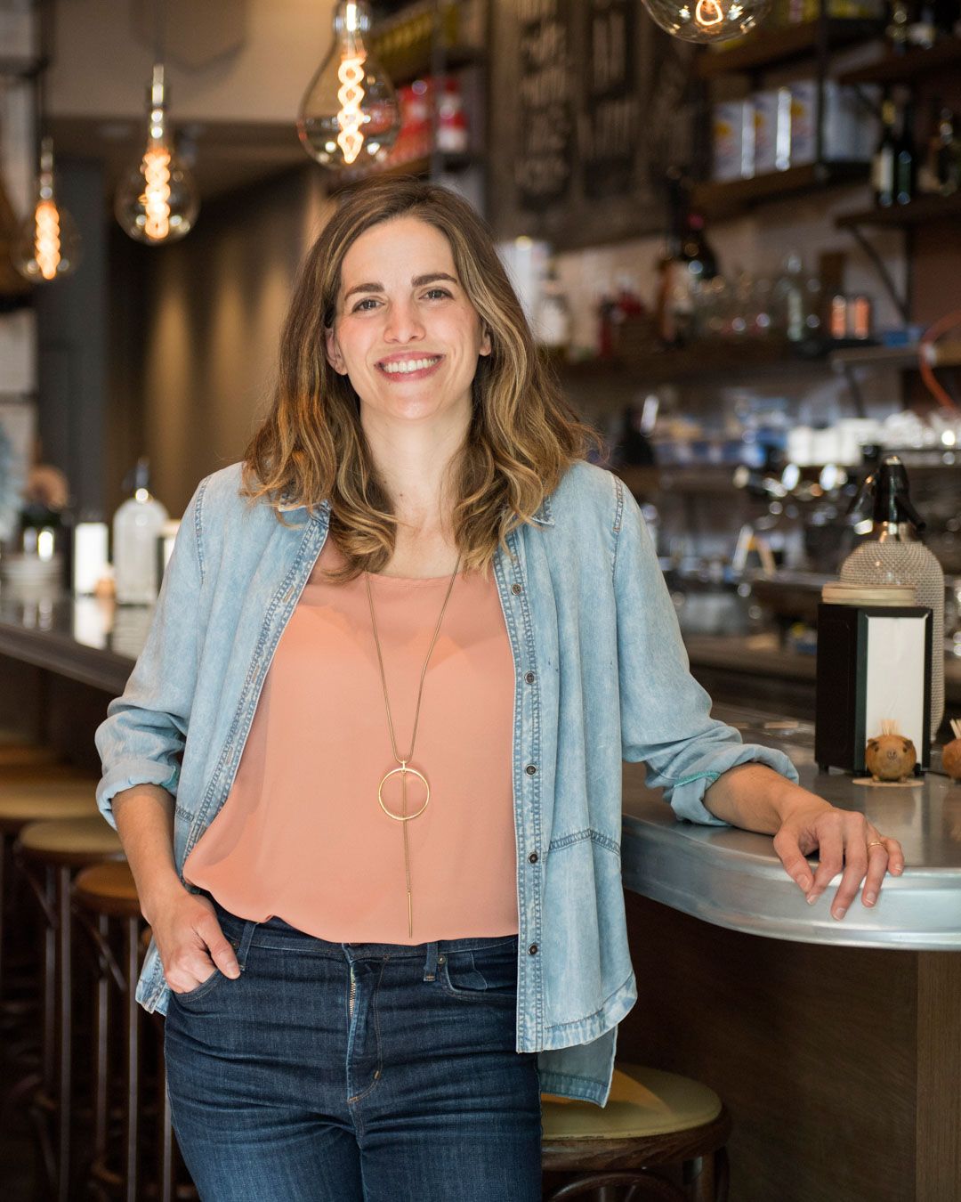 Katie Button smiles at the camera in a restaurant and bar by the bar counter.