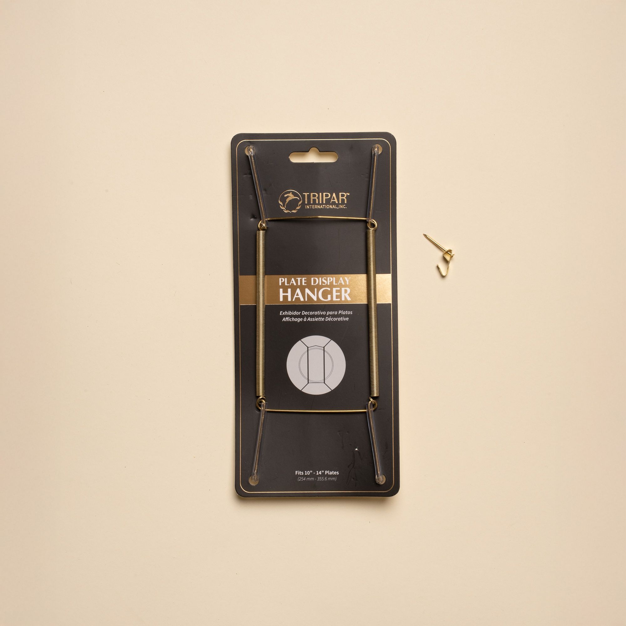 A brass plate hanger in its black cardboard package, with a nail off to the side.
