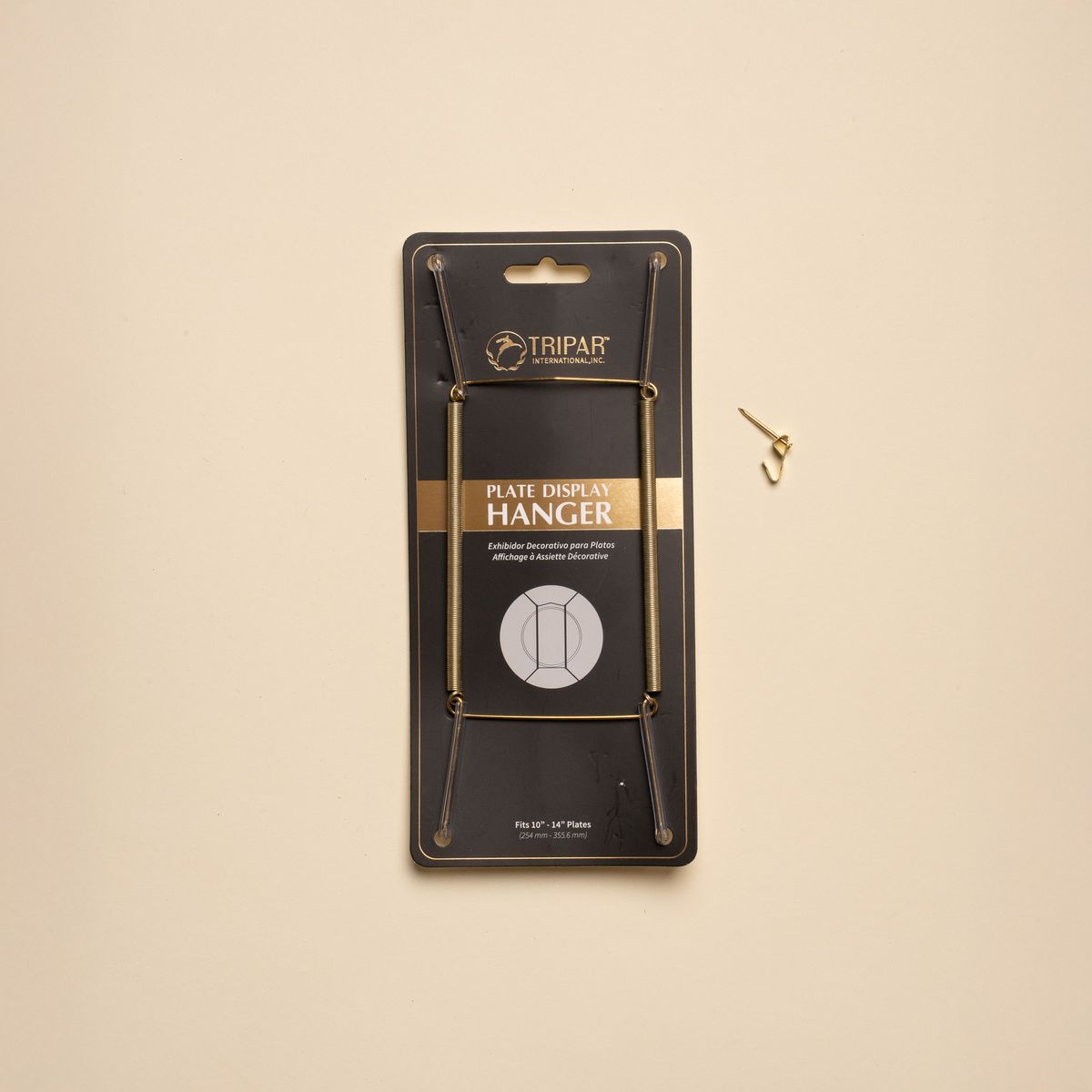 A brass plate hanger in its black cardboard package, with a nail off to the side.