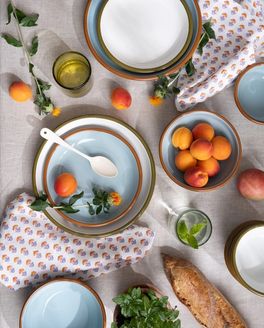 An overhead view of enamel plates and bowls in light blue and white colors with floral block printed napkins, enamel spoon, glassware, and foods.