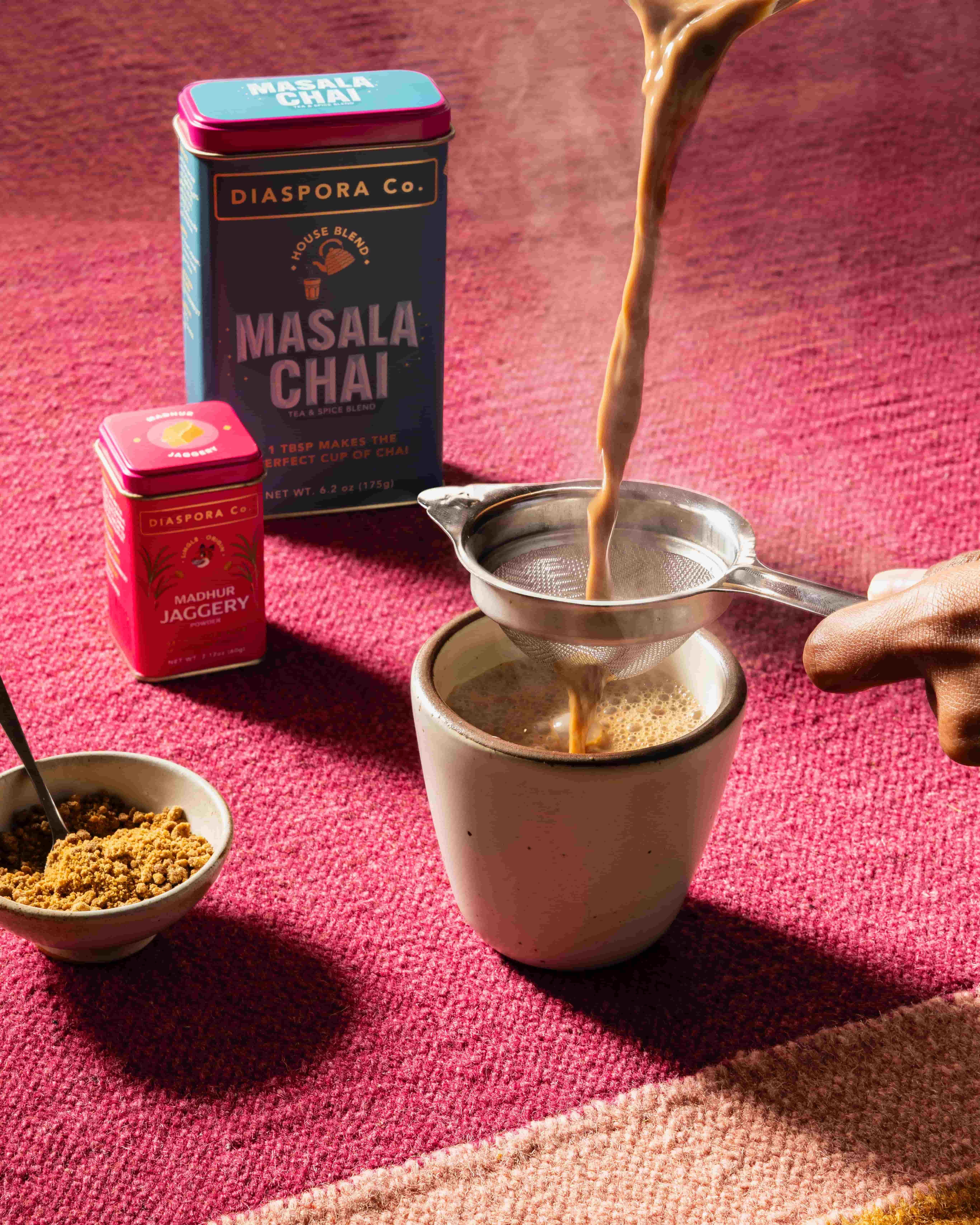 A hand holds a strainer over a small ceramic cup pouring chai, next to two food tins of masala chai and jaggery, a small bowl of jaggery, all against a bright pink background.