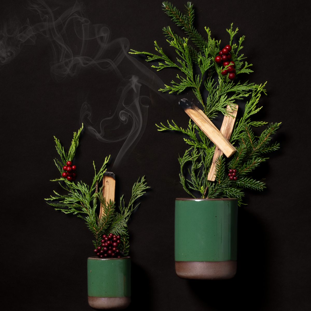 Small and large glossy emerald green ceramic candles with cedar sprigs, berries, and smoking palo santo sticks against a black background
