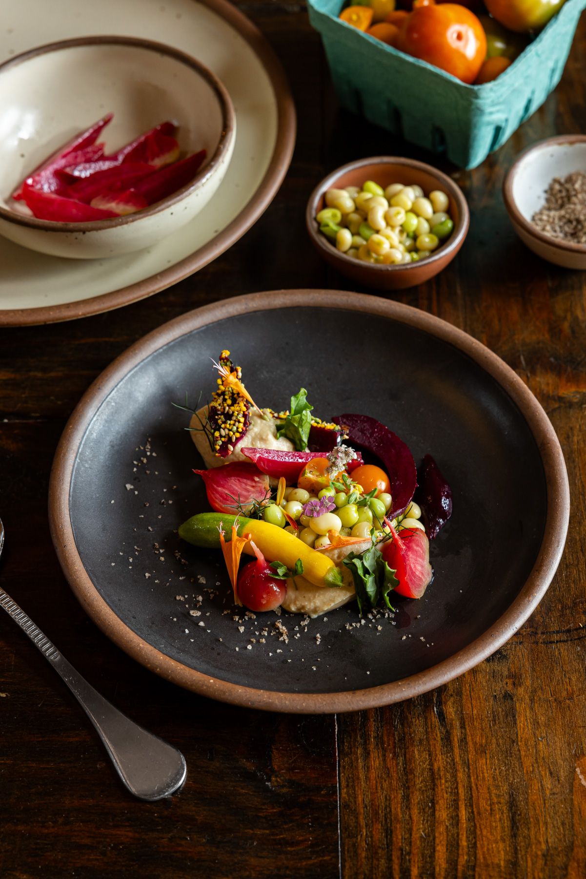 On a table is a beautiful plate of food with fresh vegetables on a ceramic black plate with other dishes and ingredients in the background.