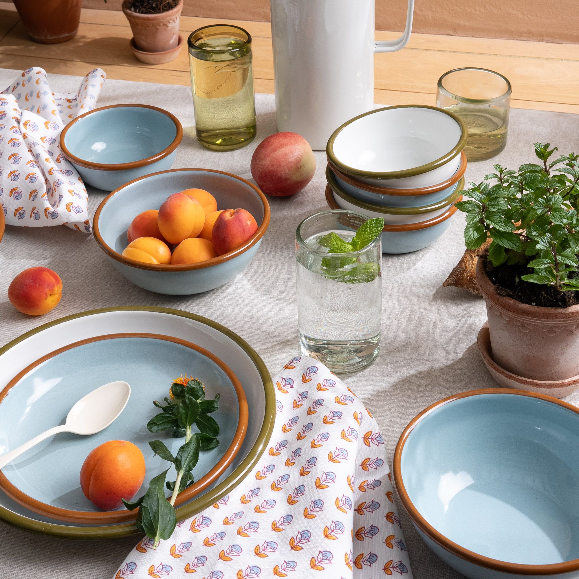 A table filled with white and robin's egg blue enamel dinnerware, with flower block printed napkins, tumbler glasses, plants and more.