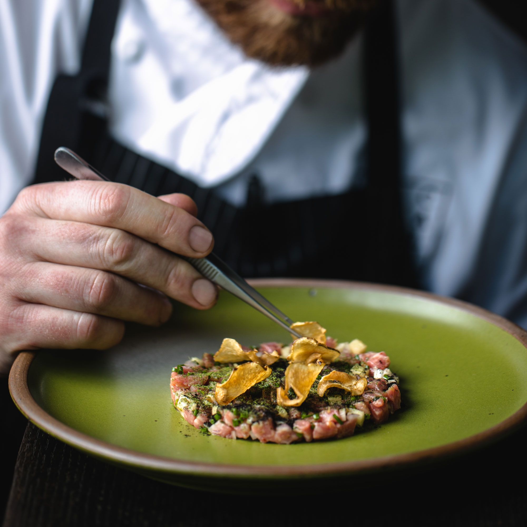 A hand holds tweezers and places a fine dish on a fern green ceramic plate