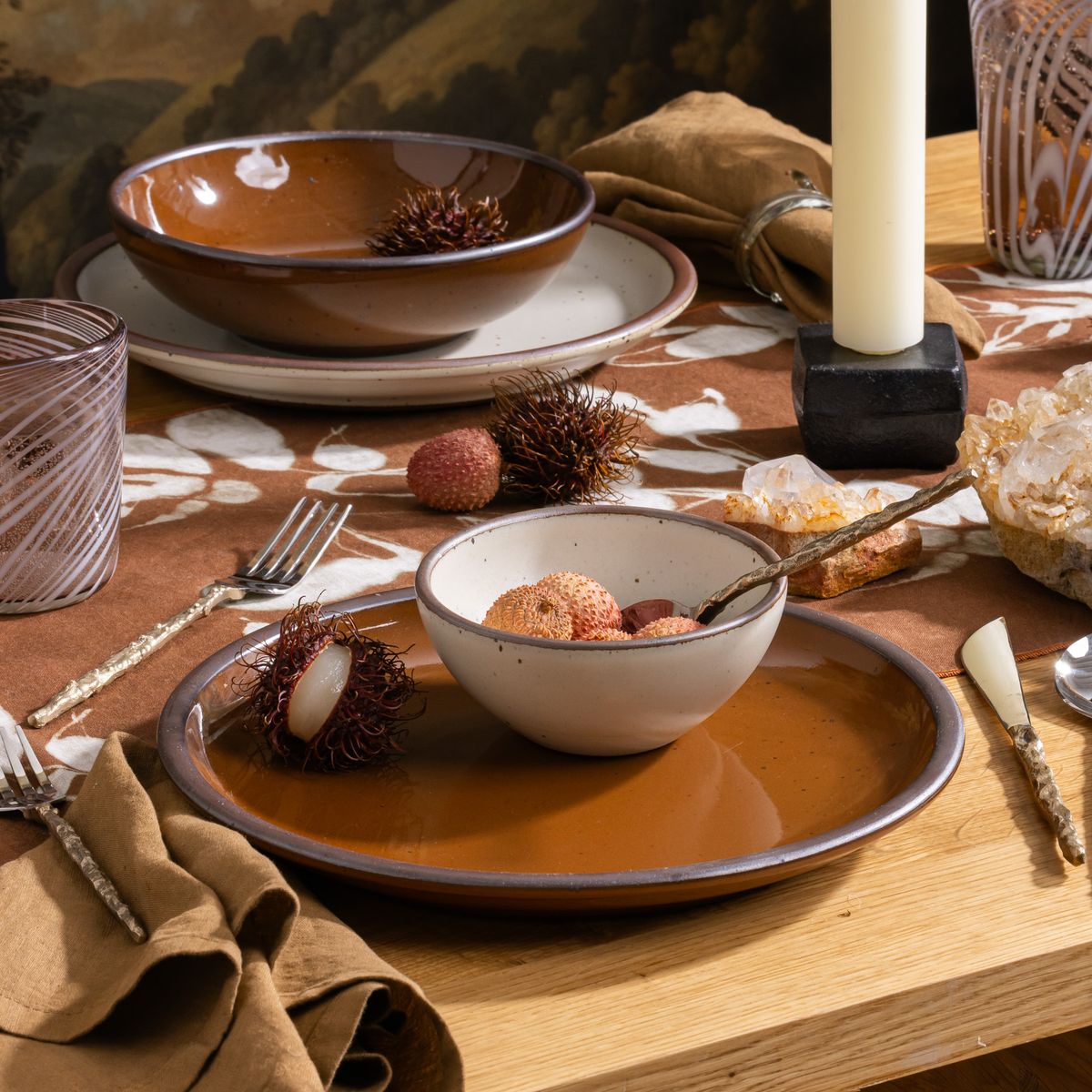Table set with glossy caramel brown dinnerware, lychee fruit, candles, and crystal accents on a patterned brown table runner.