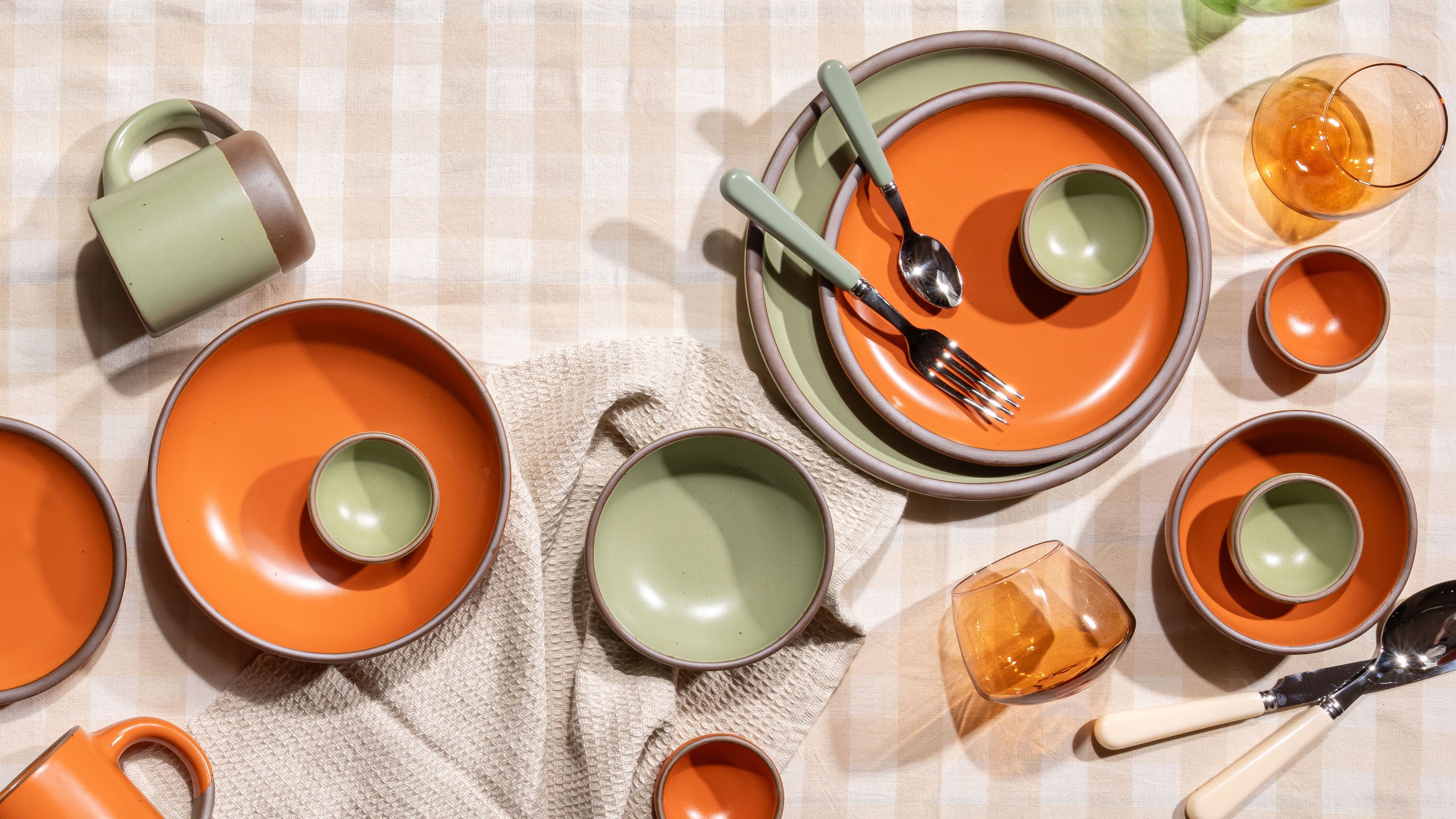 An overhead view of ceramic plates and bowls artfully arranged in bold orange and sage green colors, surrounded by flatware glasses and more. All against a natural gingham tablecloth.