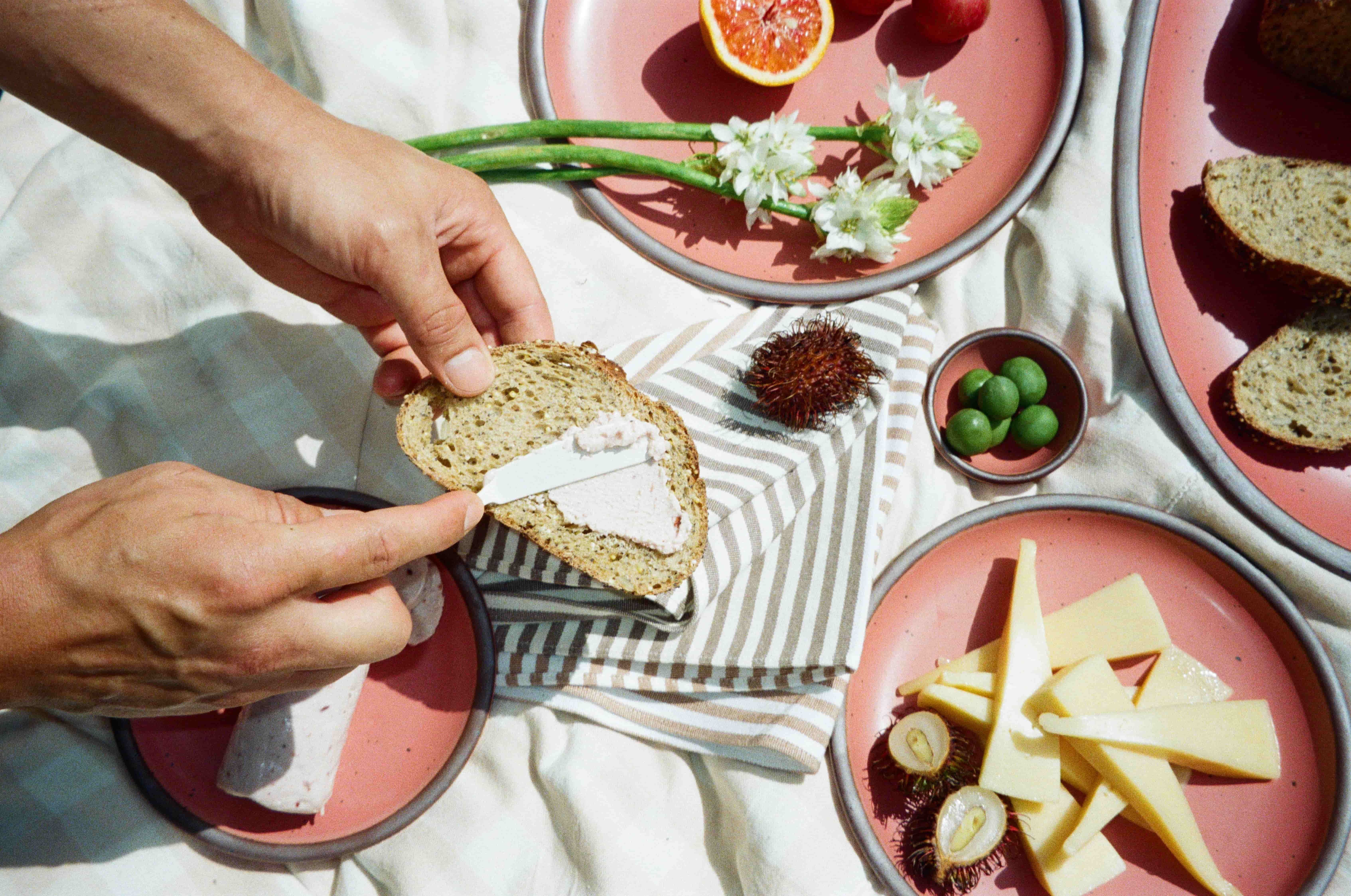 Spreading pâté on seeded bread with cheese, fruit, and fresh herbs arranged on watermelon pink ceramic plates outdoors.