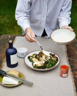 A person dresses sauces over roasted broccoli on an outdoor table. Also on the table is a bottle of olive oil, seasonings, and parmesan