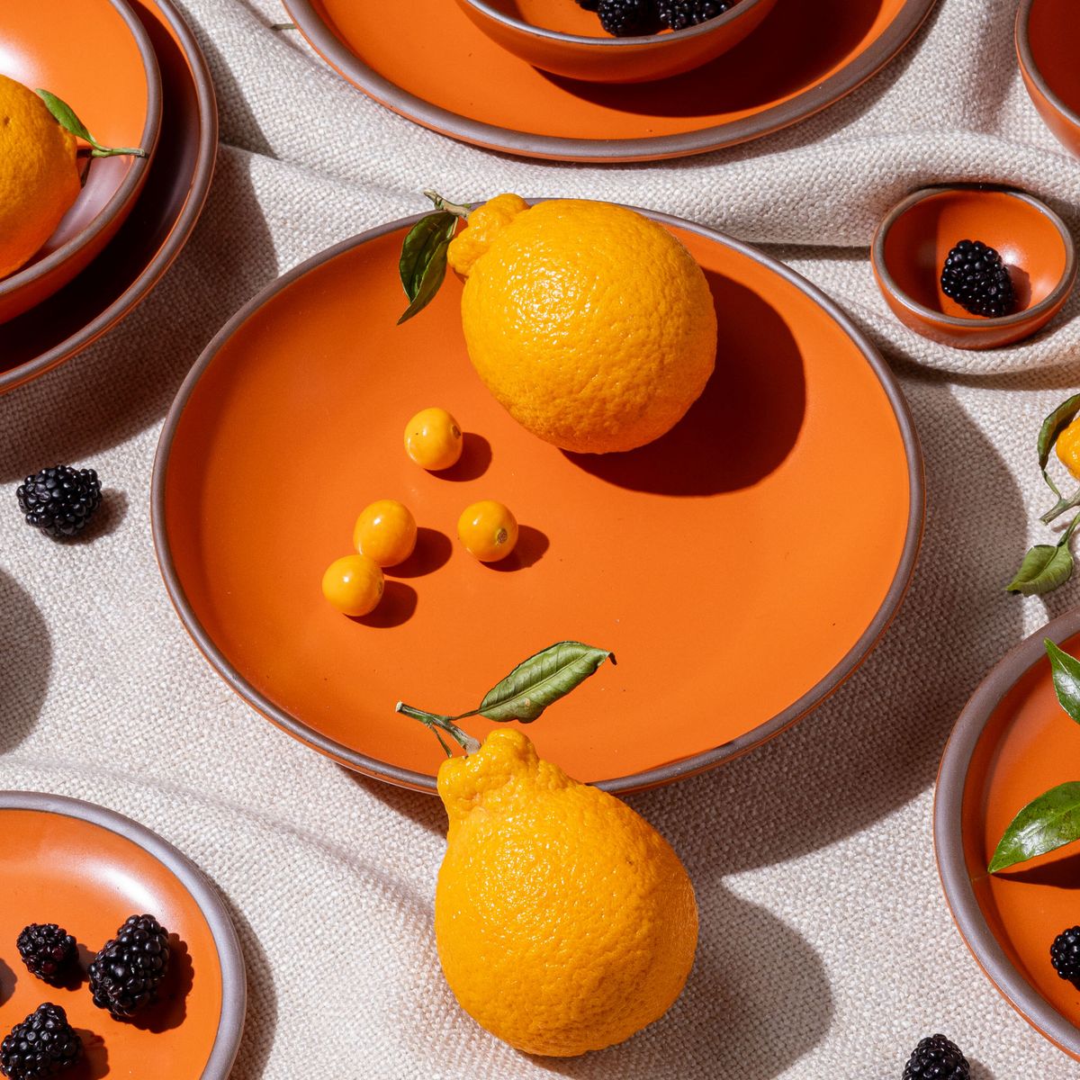 A large ceramic plate with a curved bowl edge in a bold orange color featuring iron speckles and an unglazed rim, topped with oranges and surrounded by other bowls and plates.