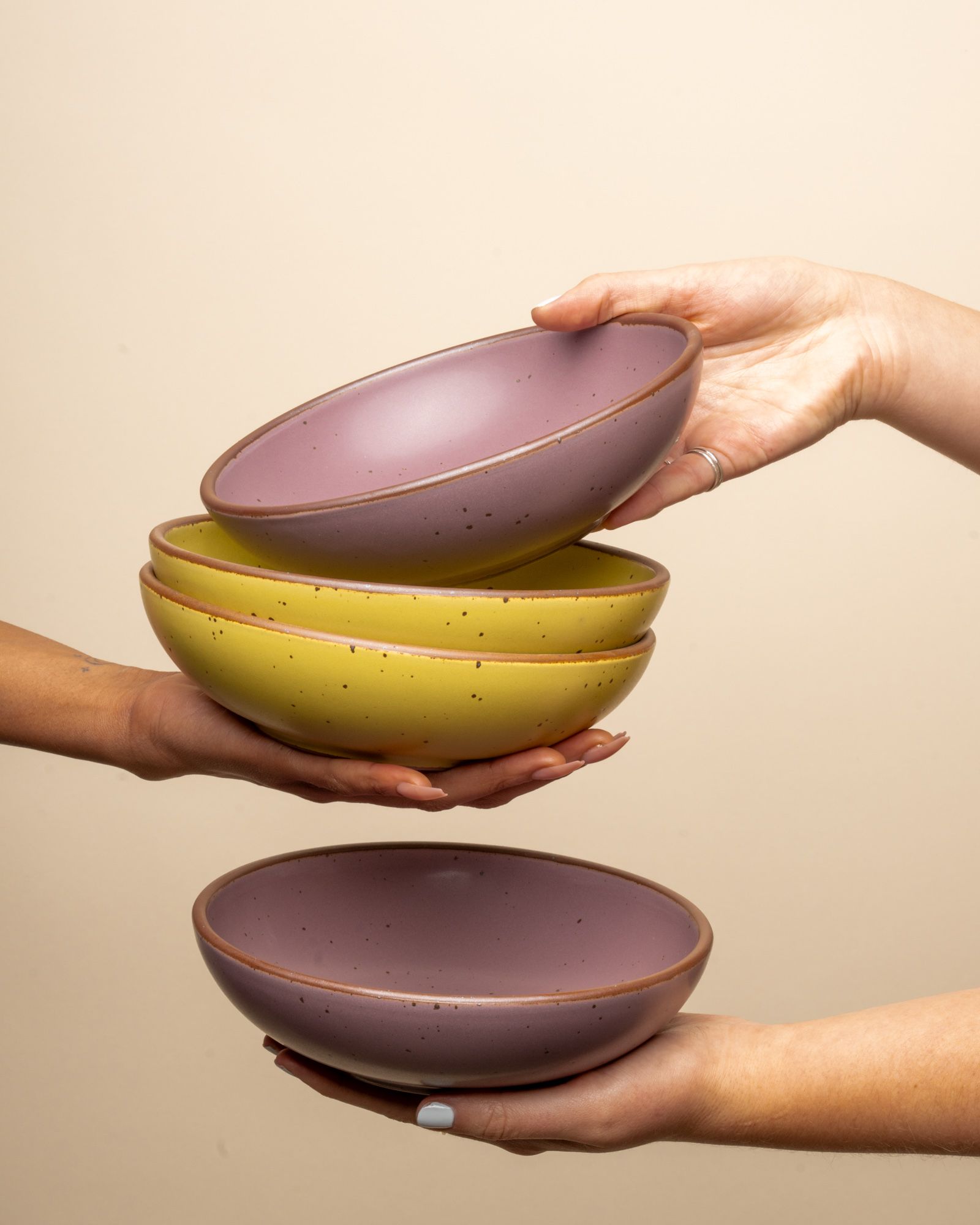 Hands exchanging shallow ceramic bowls in zesty yellow and soft mauve purple colors.