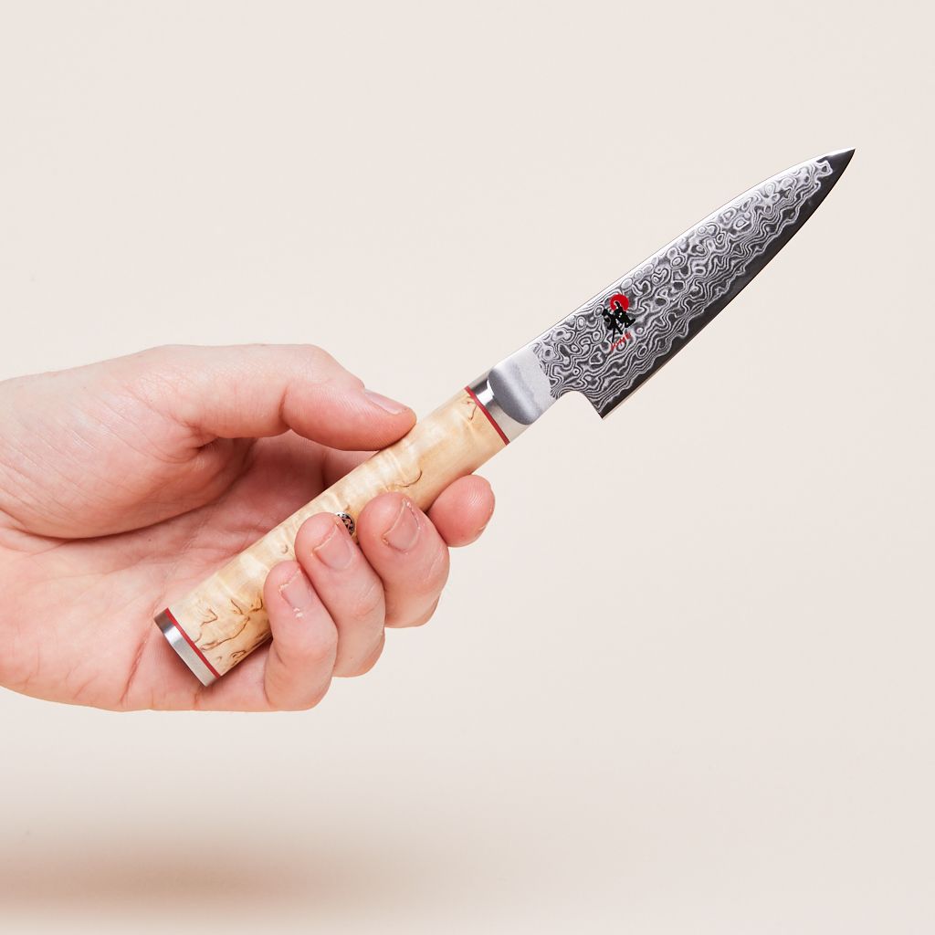 Fingers grip the birchwood handle of a paring knife with a metal blade.