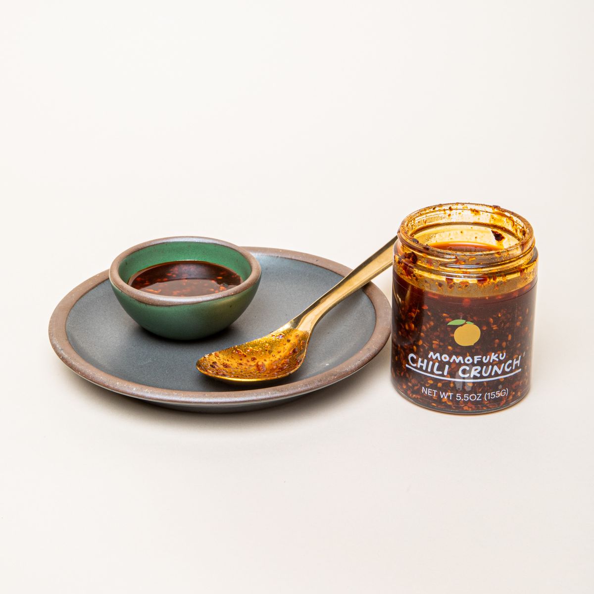 An open clear jar labeled "Momofuku Chili Crunch" filled with a warm brown sauce with chili flakes next to a plate, tiny bowl, and brass spoon