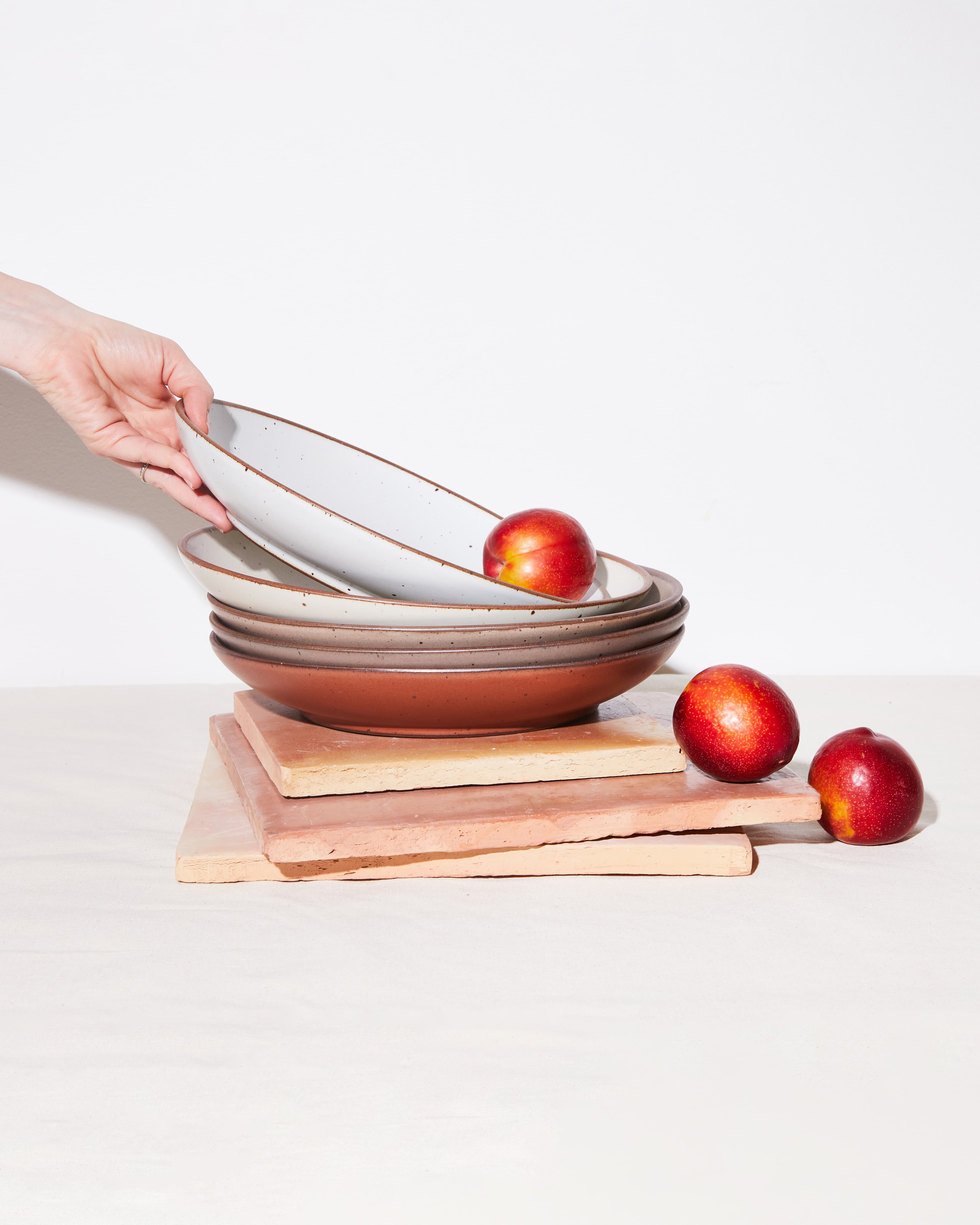 A stack of ceramic coupe plates in terracotta, tan, and white colors with apples falling out of them