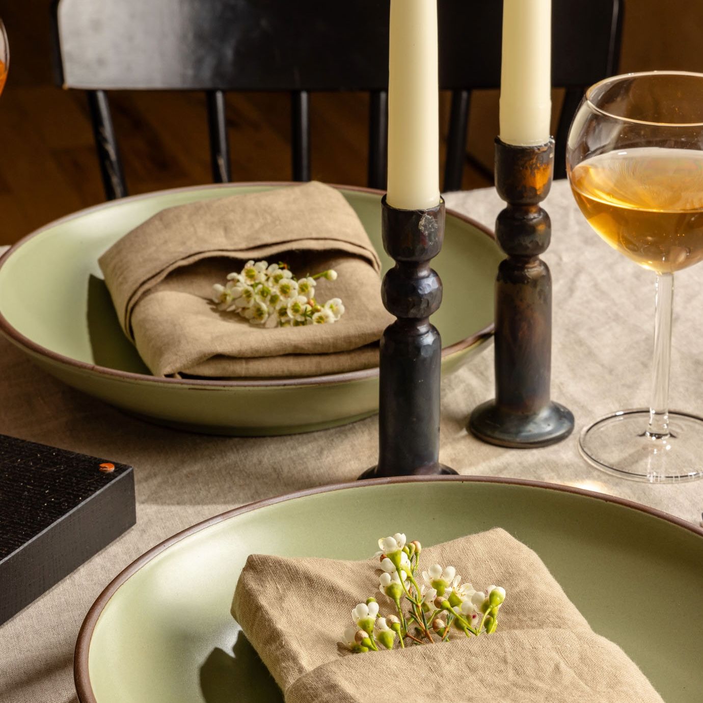Iron candleholders with cream taper candles sit on a dinner table surrounded by two plates in a sage green, folded napkins with delicate flowers, and a glass of wine.