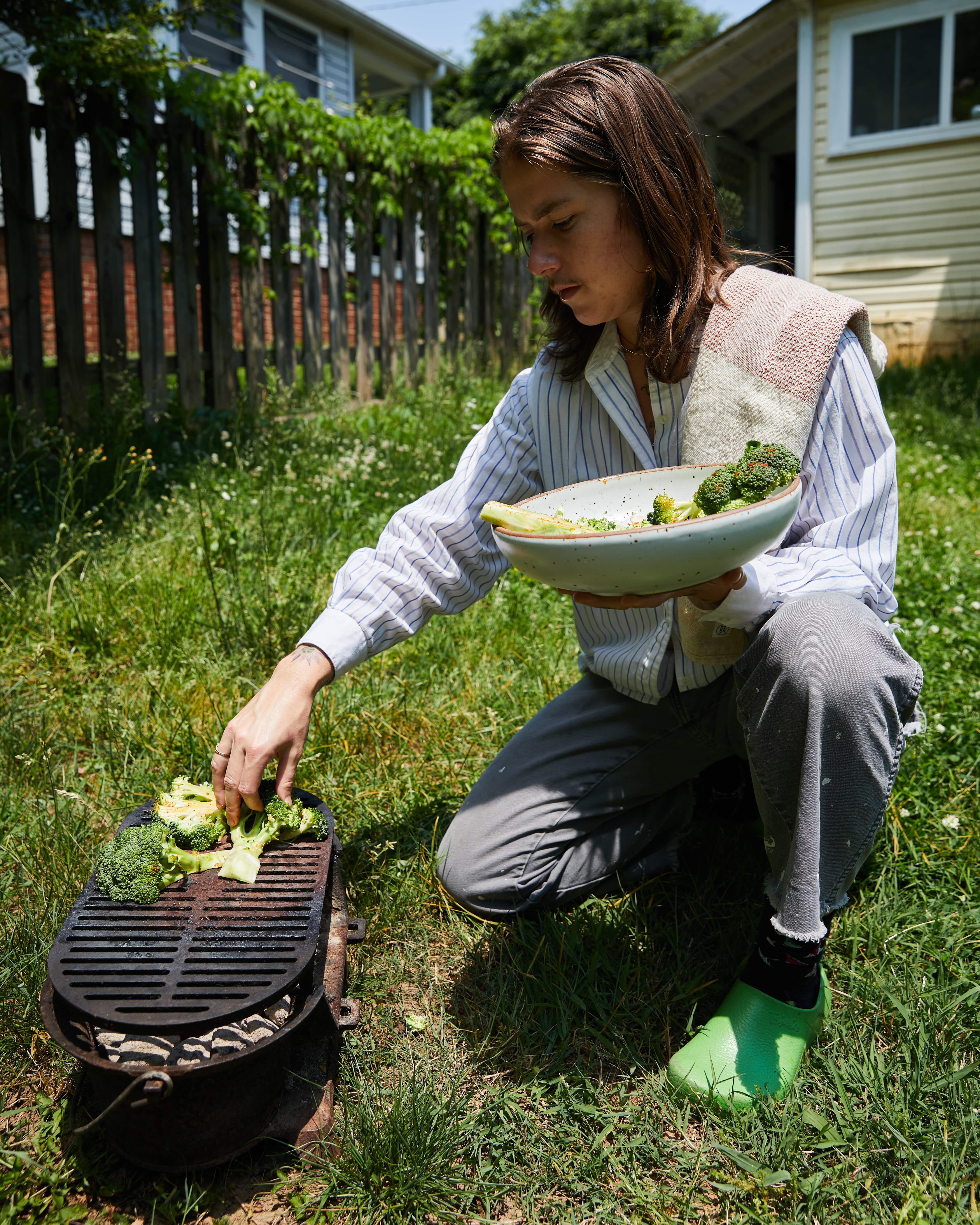 Outside, A person is taking charred broccoli off a grill and putting it into a bowl