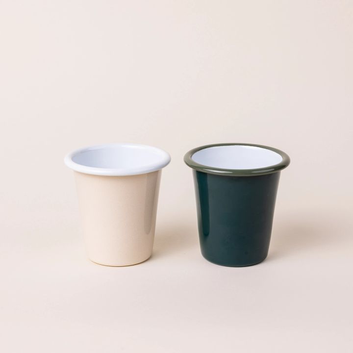 Two short enamel cups next to each other, the left with a cream exterior and white interior, and the right with a dark green teal exterior and white interior