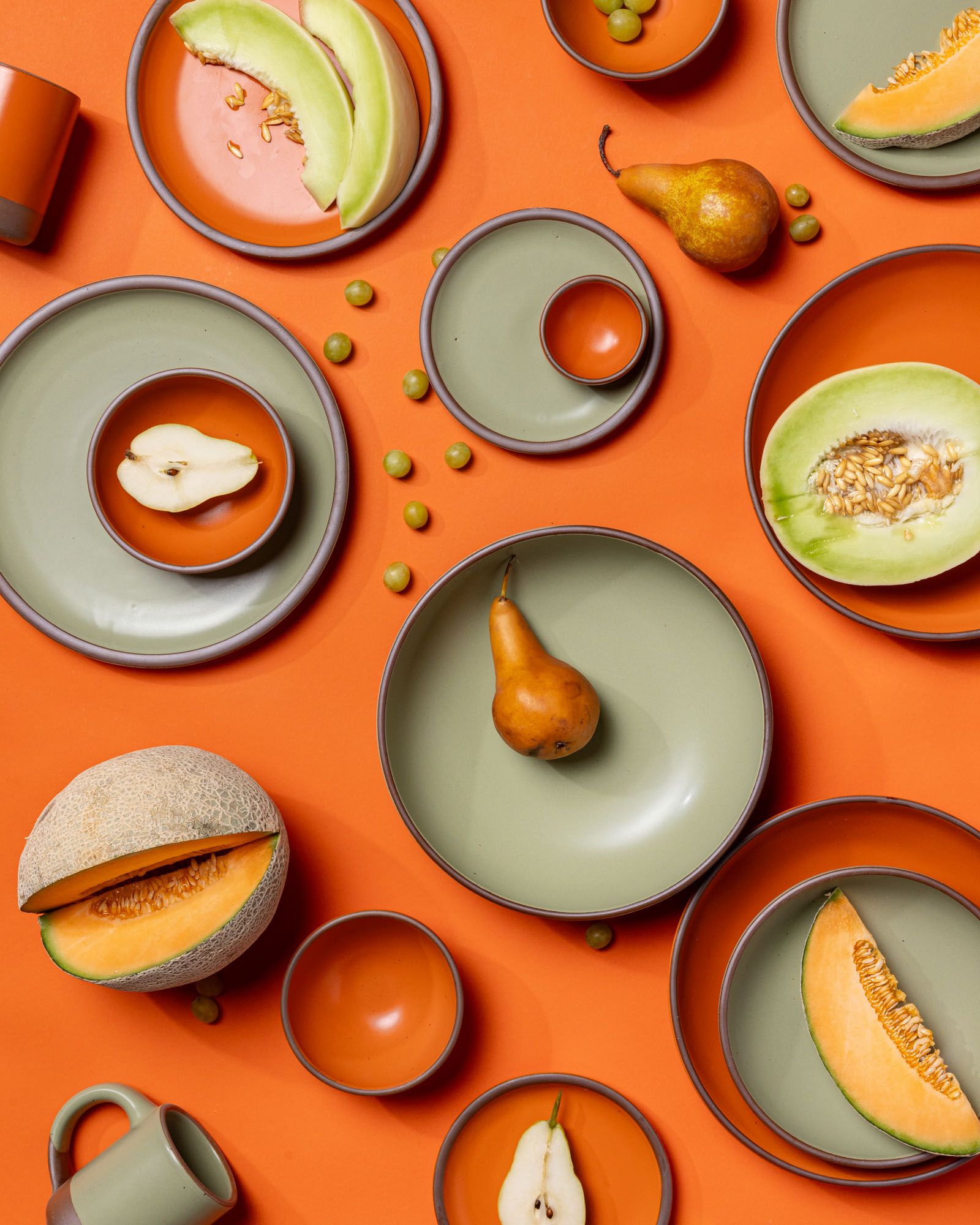 Ceramic plates and bowls in sage green and bold orange are artfully arranged together with different fruits in similar colors against a bold orange background.