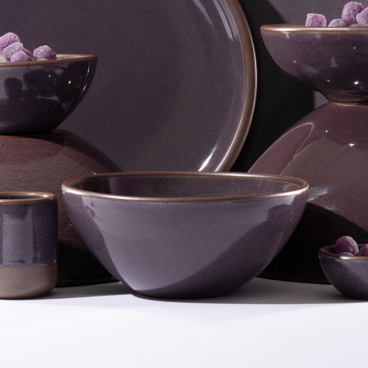 A ceramic soup bowl in a purple color with a glowing sheen surrounded by other ceramic dinnerware pieces.