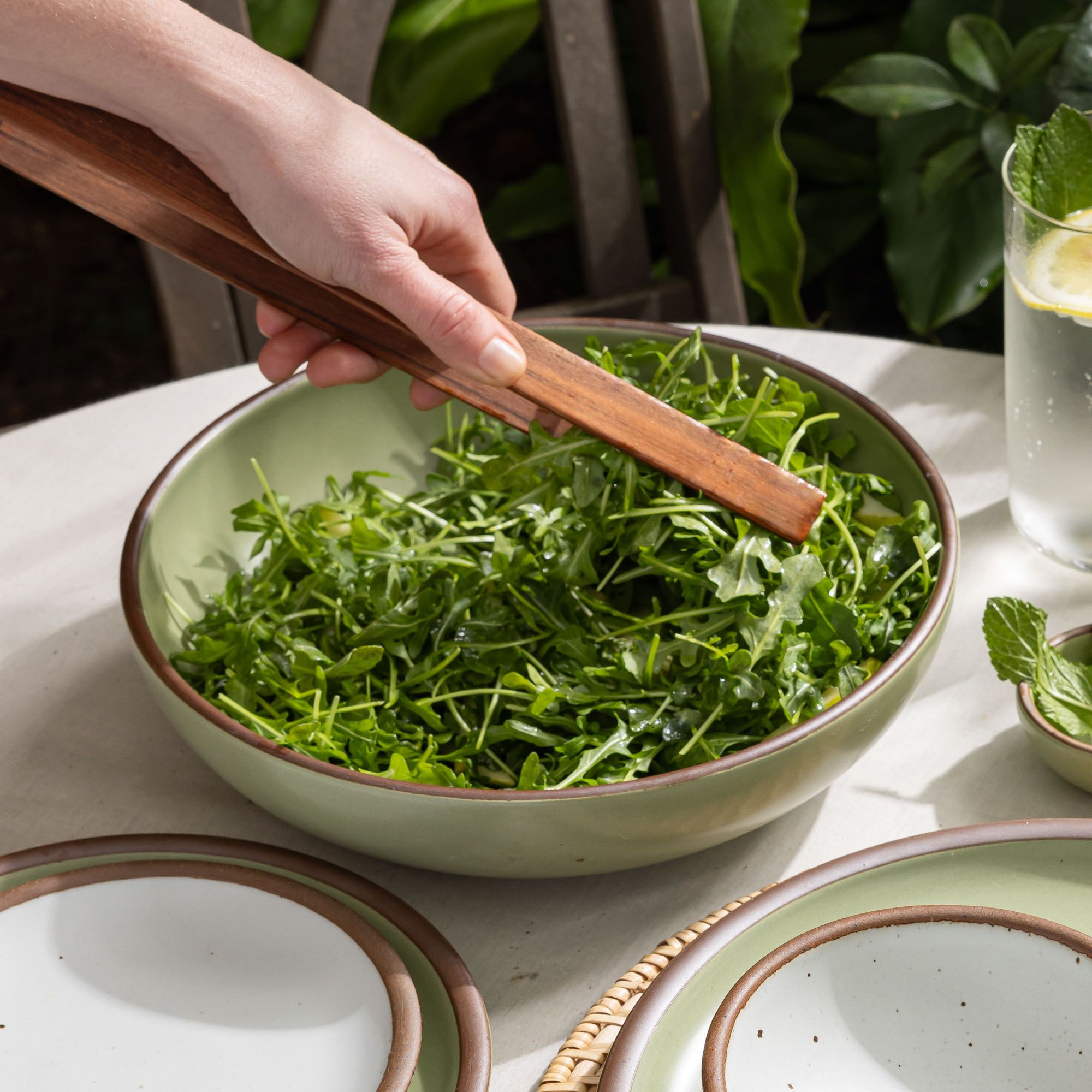 A salad sits in a large shallow serving ceramic bowl in a calming sage green color with a hand holding serving tongs.