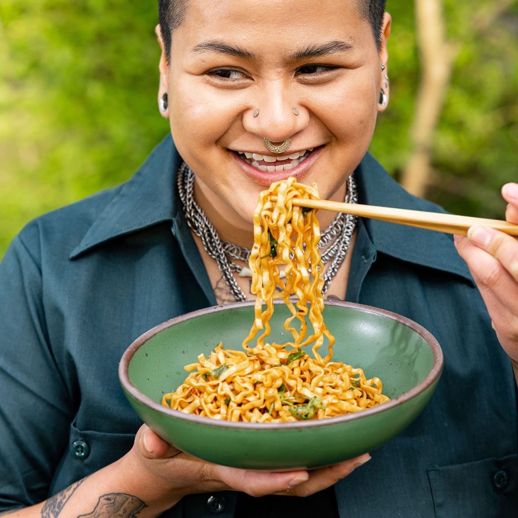 A person is smiling while eating noodles out of a shallow ceramic bowl with chopsticks.