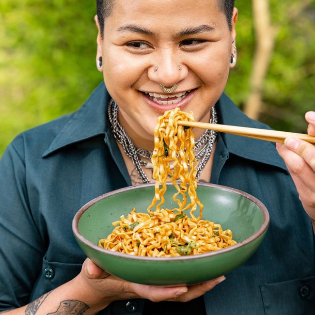 A person is smiling while eating noodles out of a shallow ceramic bowl with chopsticks.