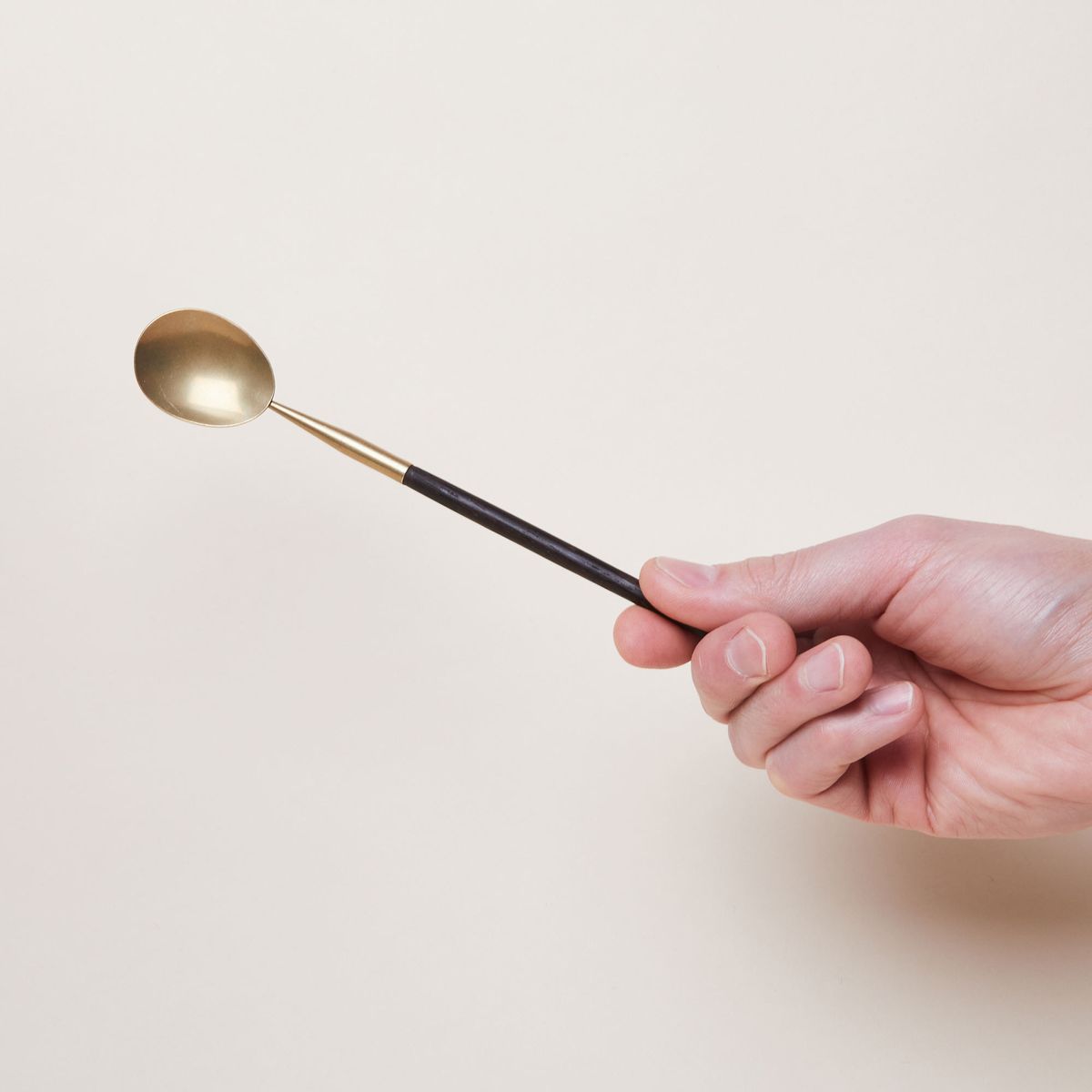 A hand holds a long spoon that has a thin, dark handle attached by a brass ferrule and bowl