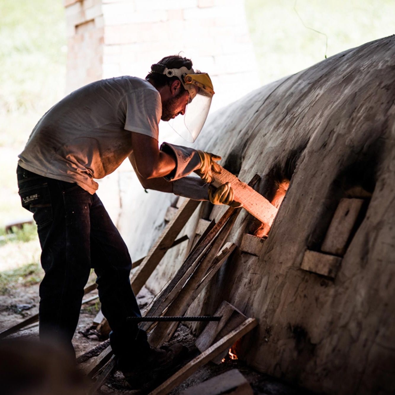 Alex putting wood into the window of an outdoor wood-fired kiln.