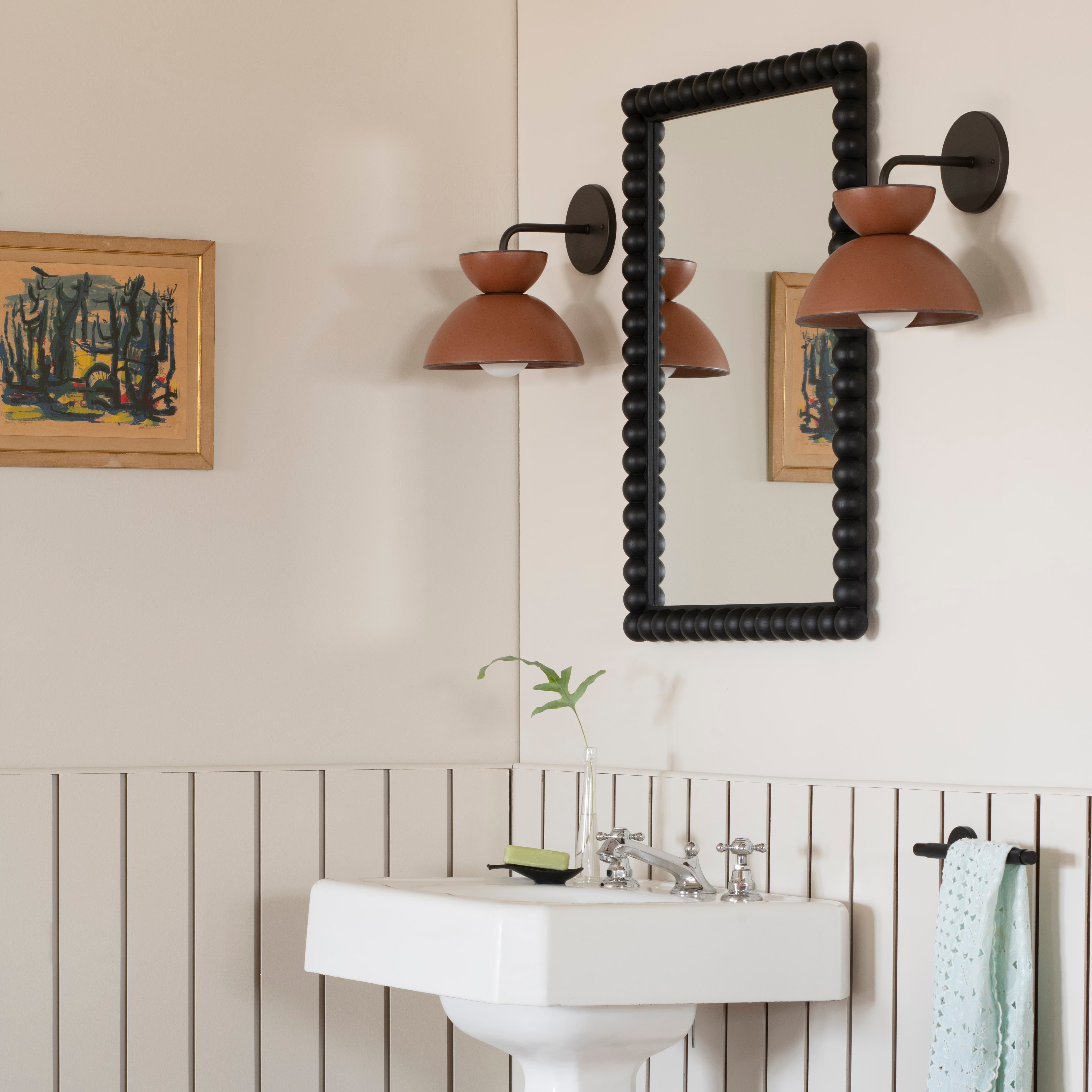 Two light fixtures made from amaro East Fork bowls surround a mirror in a black frame above a pedestal sink 