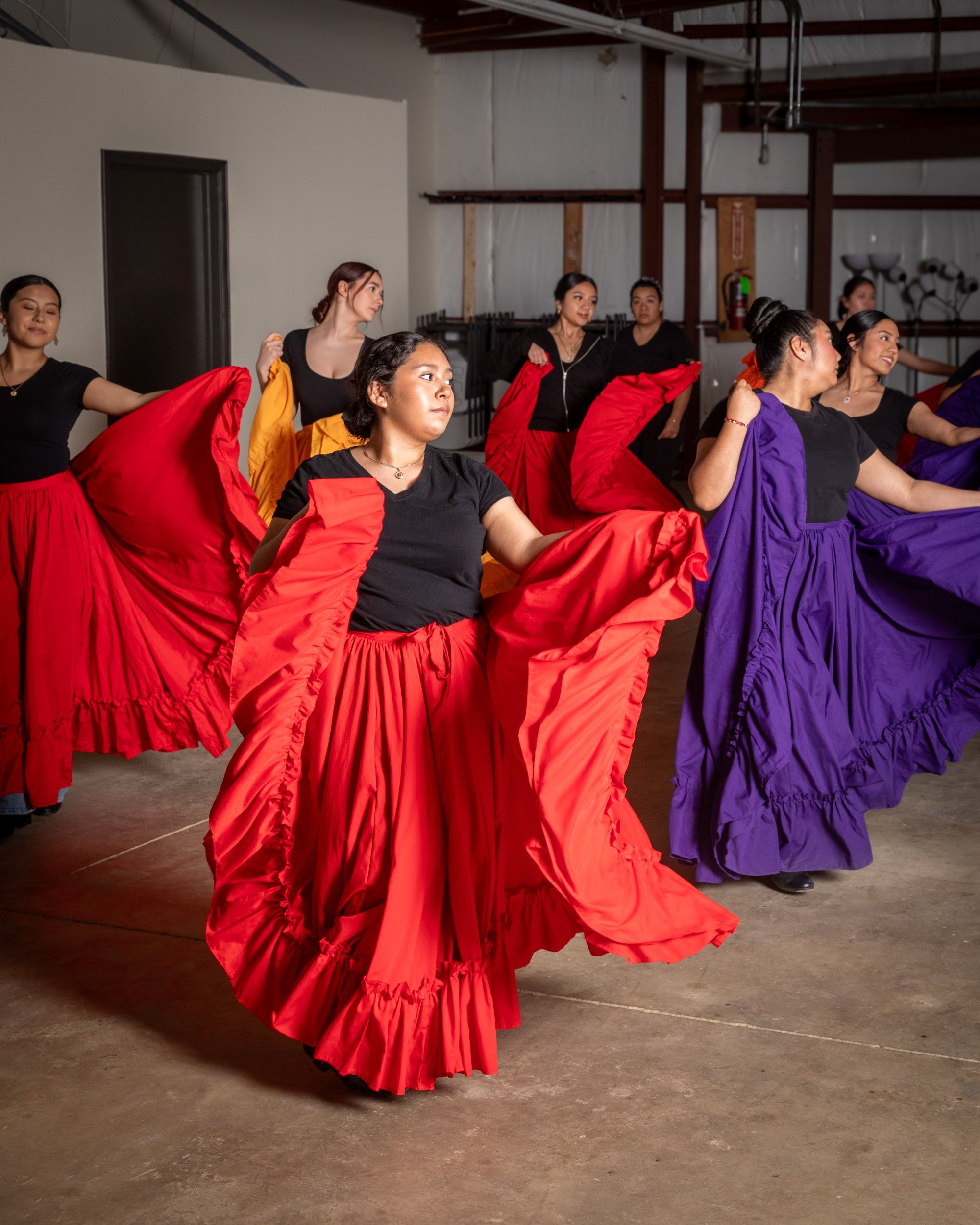 In a rehearsal space, there are young girls dancing with large colorful long skirts in red, purple, and orange colors.