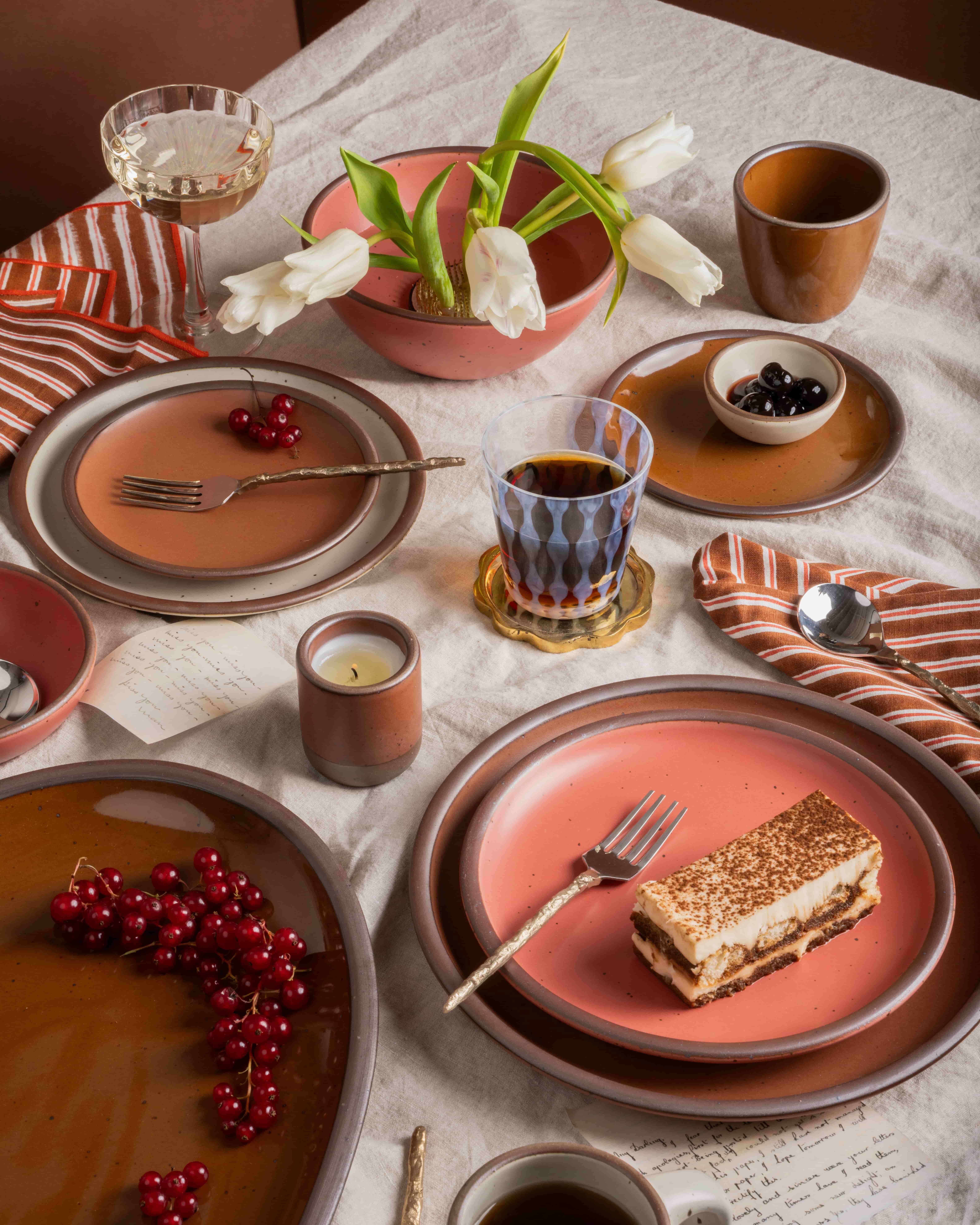 Table setting with ceramic plates in cool terracotta, watermelon pink and glossy brown, striped napkins, tulips in a bowl, drinks, and dessert on a linen tablecloth