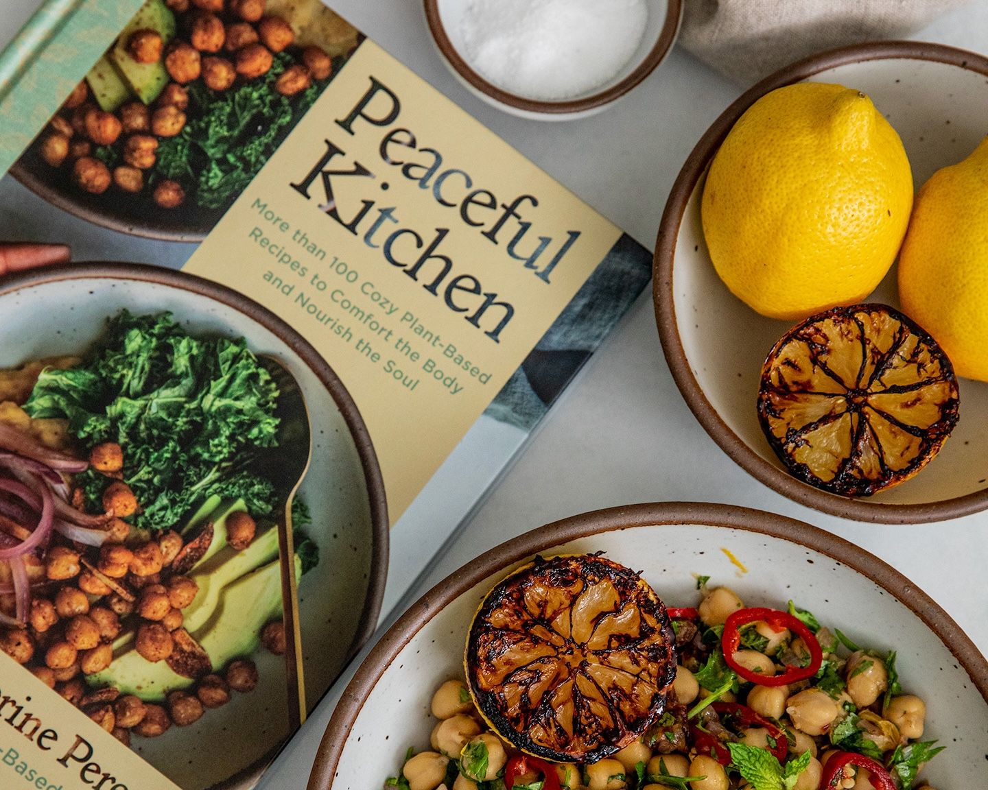 A plate filled with a chickpeas, vegetables, and lemon next to a cookbook titled, 'Peaceful Kitchen'