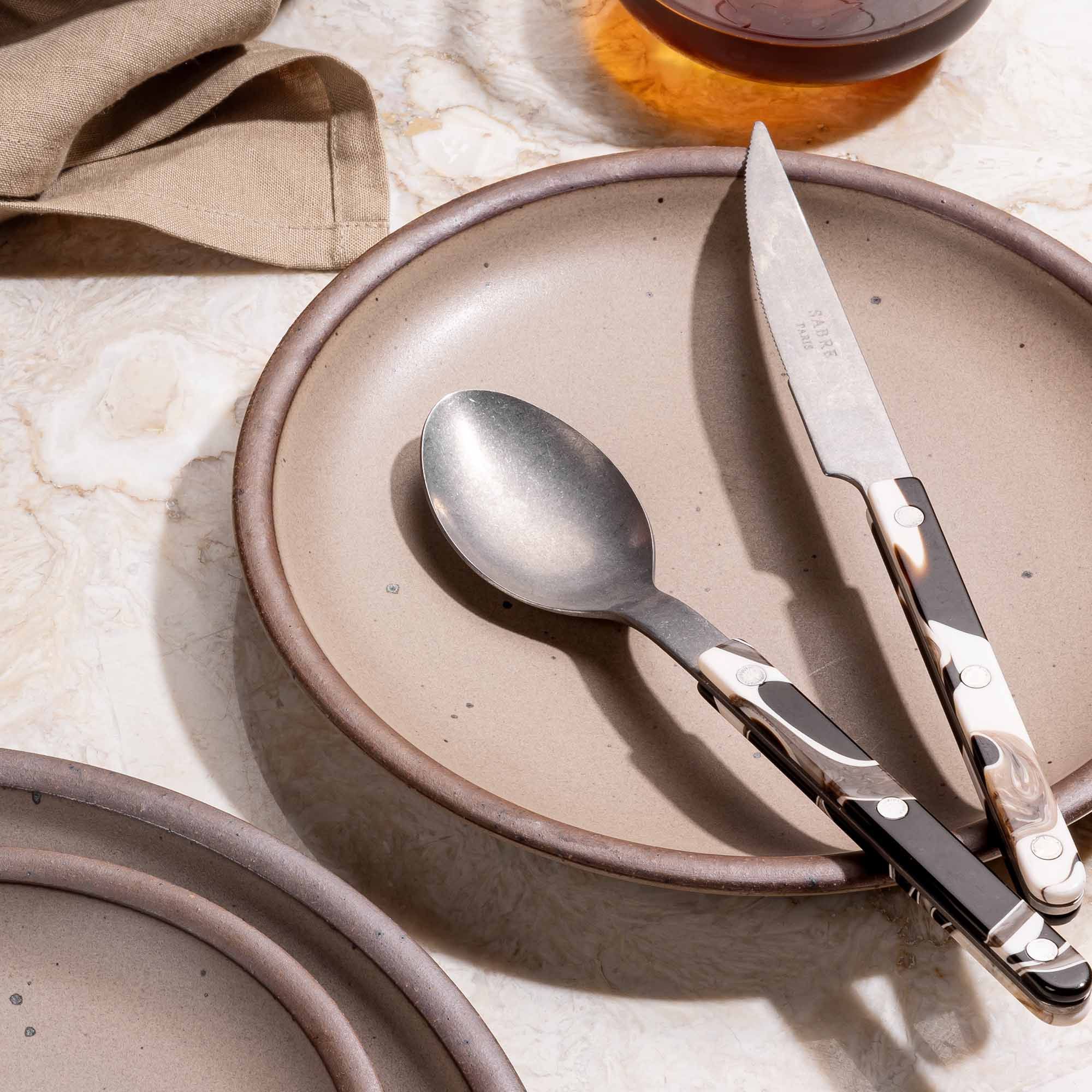 A medium sized ceramic plate in a cool tan color with marble-handled spoon and knife.