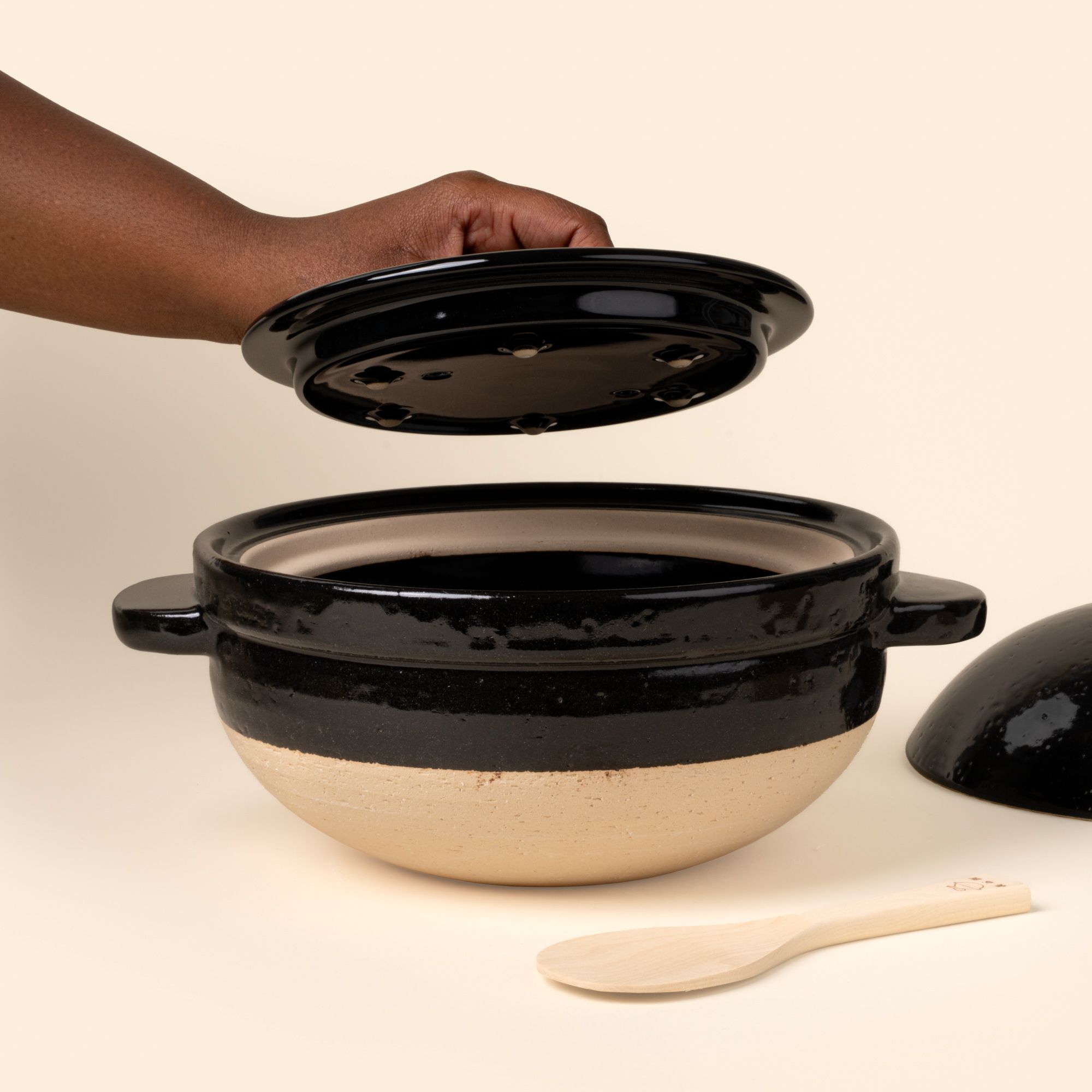 A hand the lid of a donabe rice cooker with a spoon to the side.