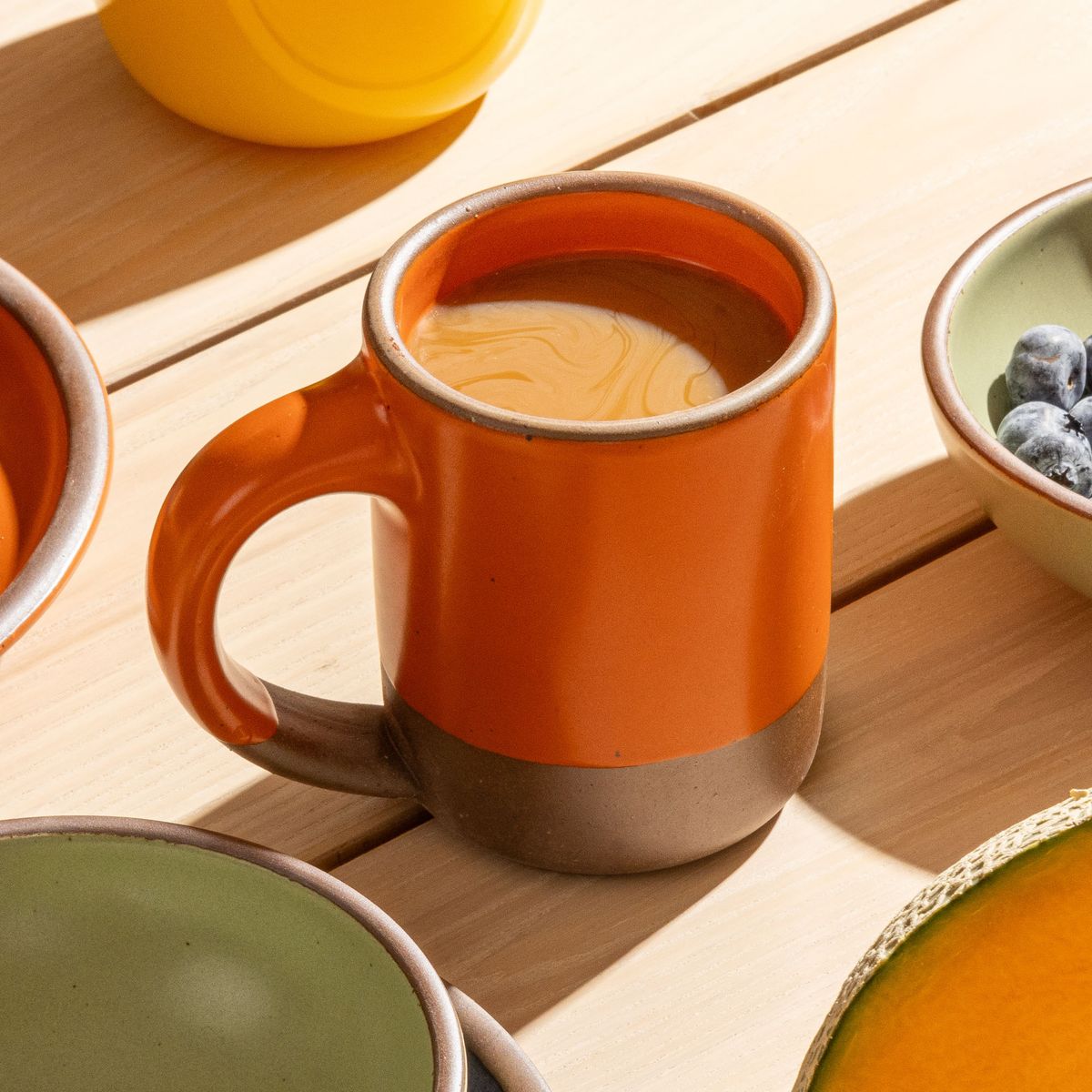 A medium sized ceramic mug with handle in a bold orange color featuring iron speckles and unglazed rim and bottom base, filled with coffee and sitting on a breakfast table