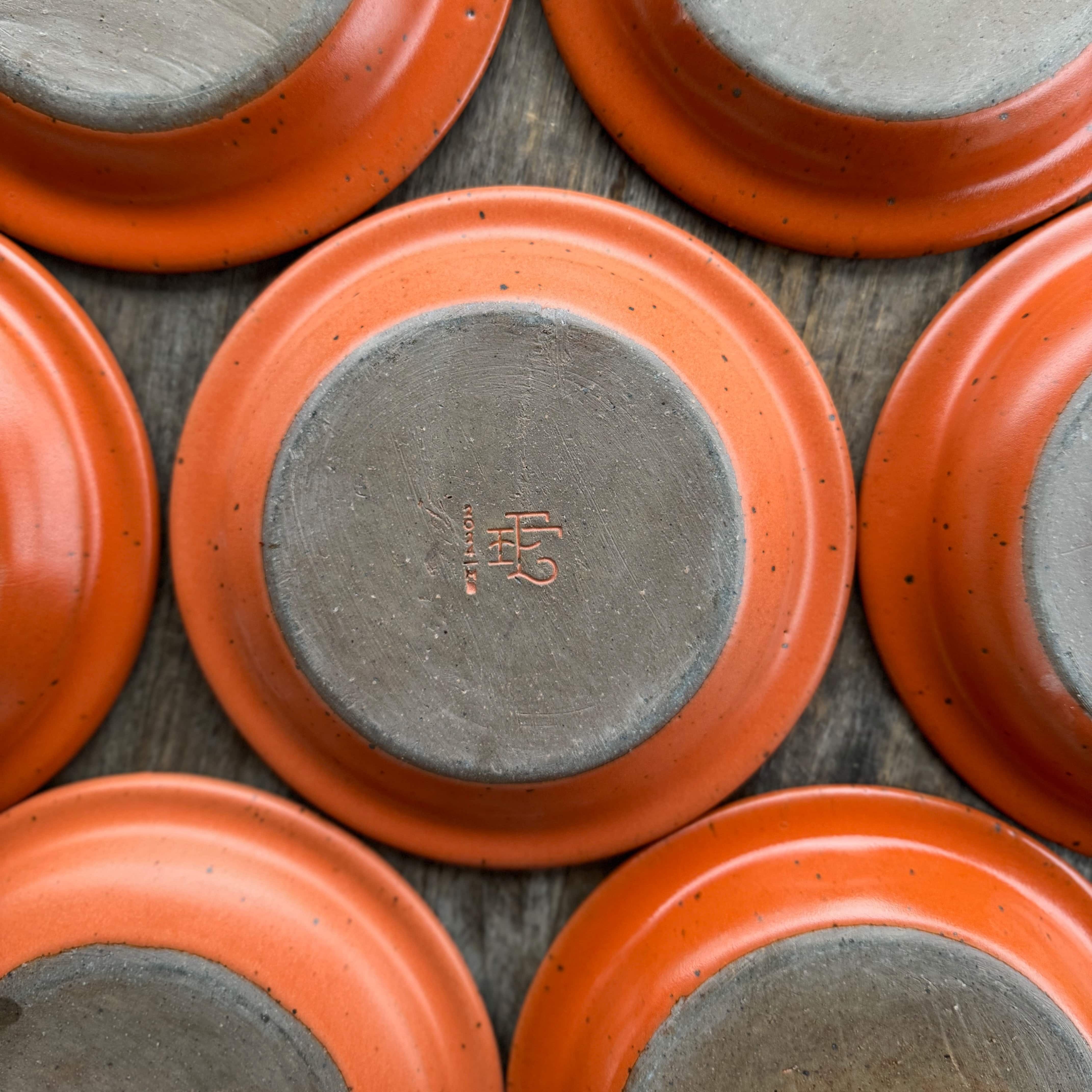 The bottom of a batch of ceramic plate-shaped incense stick holders with an unglazed rim and glazed in a bold orange color featuring lots of iron speckles. The bottom features the East Fork Workshop stamp.