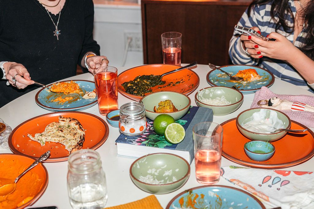 A dinner party table filled with various ceramic plates and bowls in bold colors, drinking glasses, and glimpses of people eating and talking.