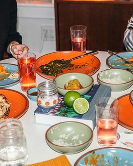 A dinner party table filled with various ceramic plates and bowls in bold colors, drinking glasses, and glimpses of people eating and talking.