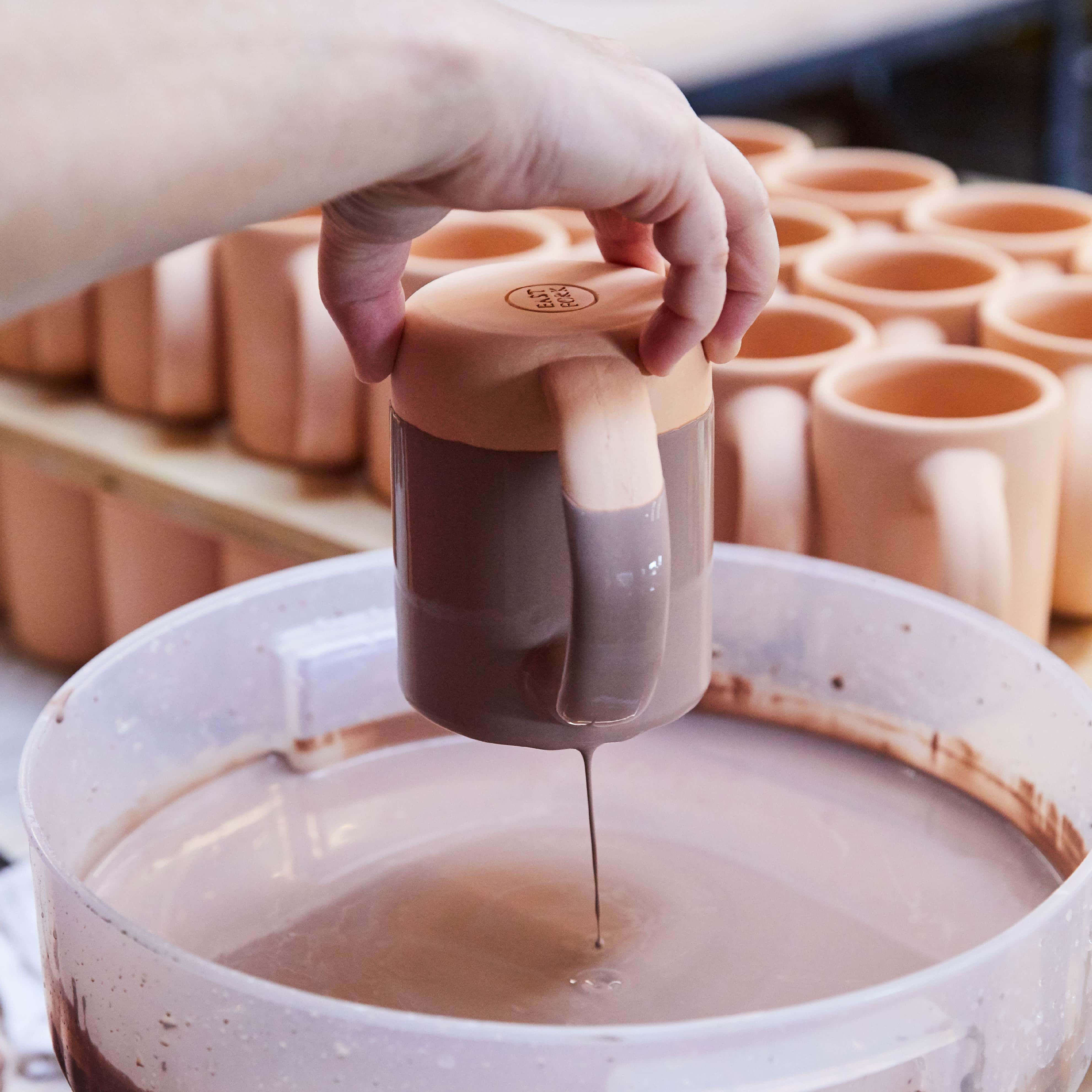 A hand holds a mug upside down dipping it into a bucket of glaze. Part of the excess glaze drips off the bottom.