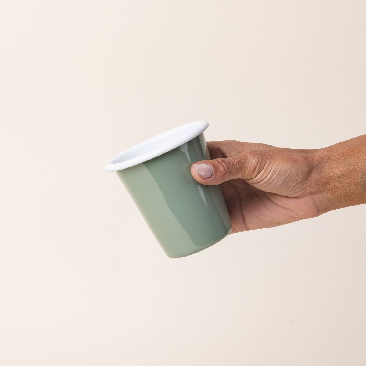 A hand holds a short enamel cup with a sage green exterior and white interior