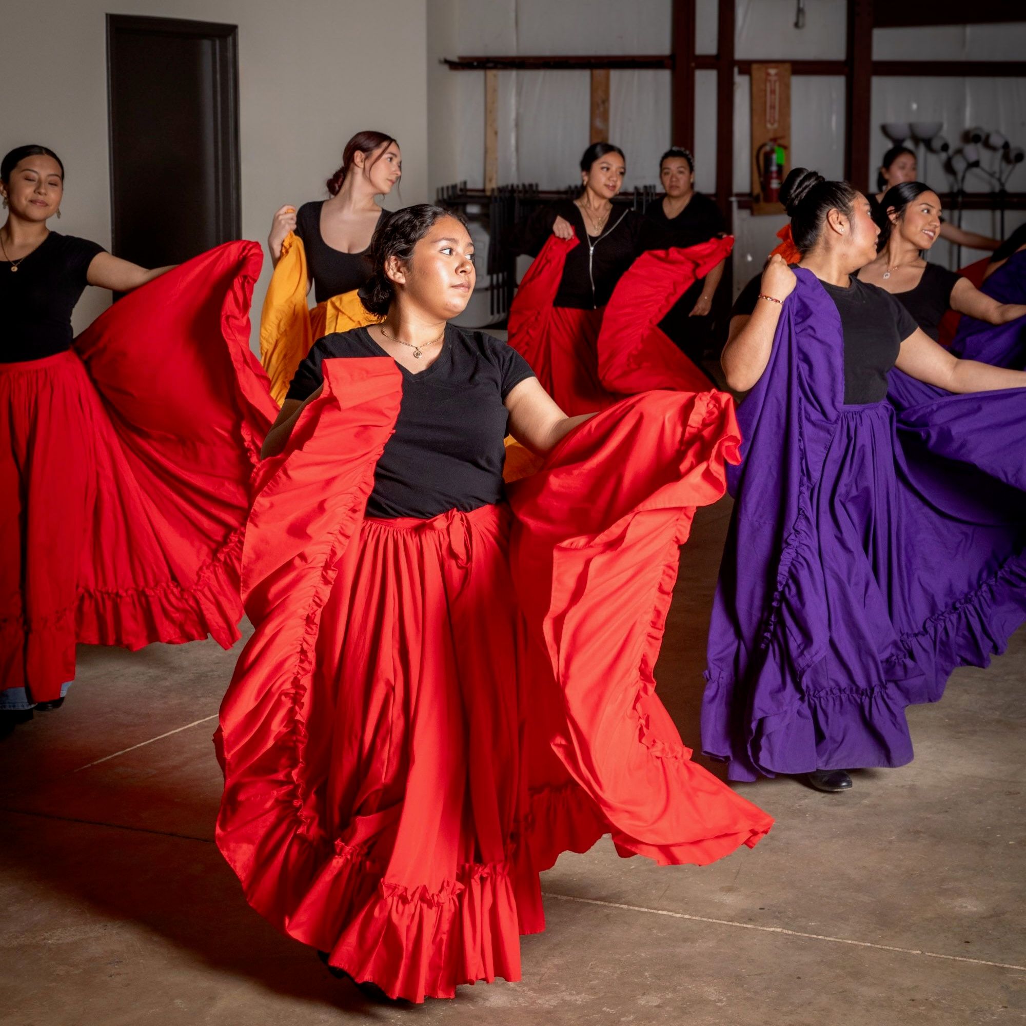 In a rehearsal space, there are young girls dancing with large colorful long skirts in red, purple, and orange colors.